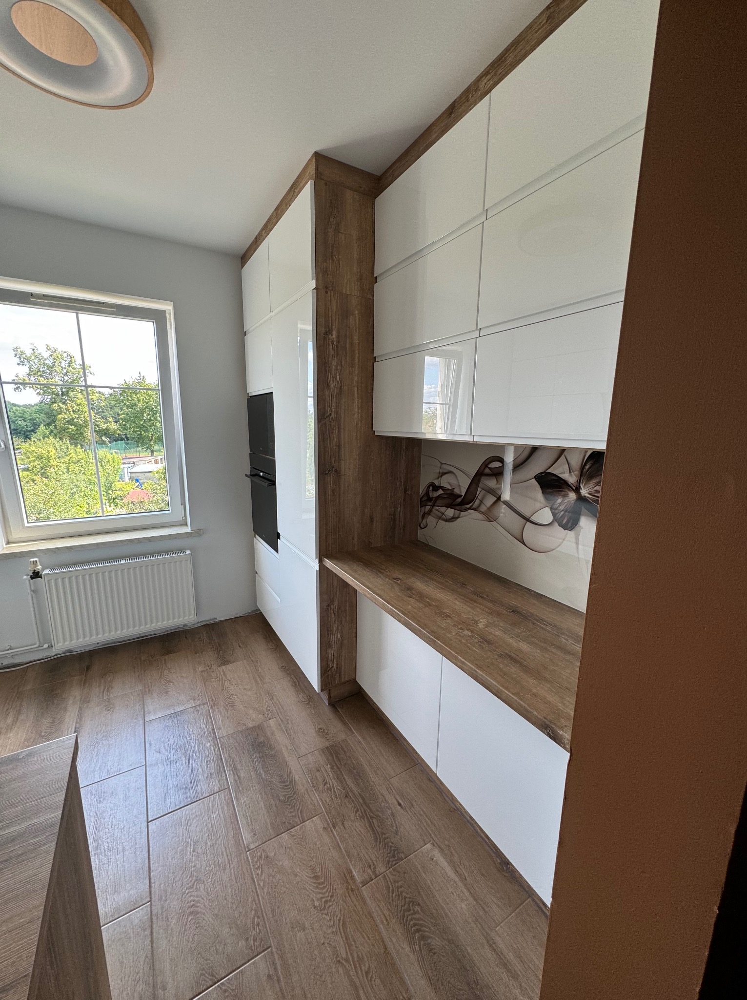 Nowoczesna kuchnia z białymi lakierowanymi frontami, blatem w kolorze drewna retro, zabudowaną piekarnikiem i płytą grzewczą, oraz dekoracyjnym panelem ściennym z motywem motyla.