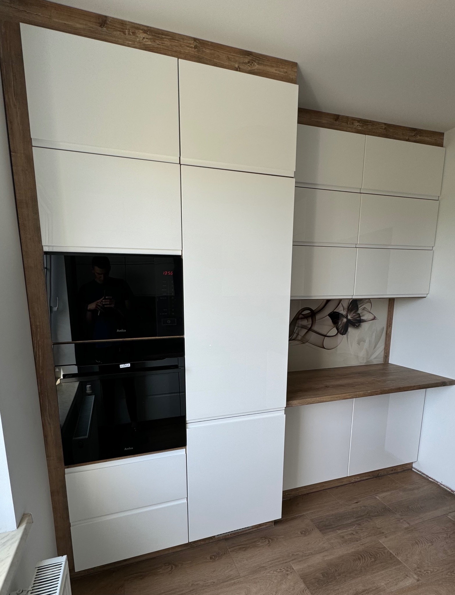 Biała zabudowa kuchenna z lakierowanymi frontami, drewnopodobnym blatem i obudową, wbudowany piekarnik i mikrofalówka Amica, widoczne płytki podłogowe imitujące drewno.