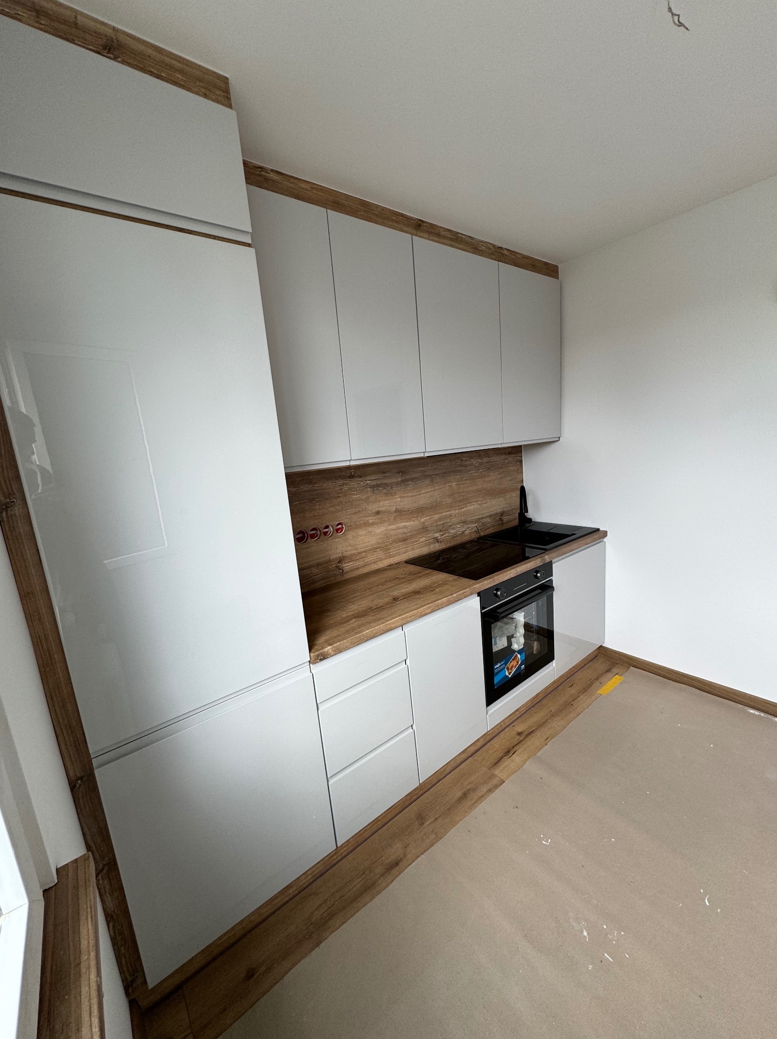 Nowoczesna kuchnia z białymi, lakierowanymi frontami i blatem w kolorze drewna retro, widoczne gniazdka elektryczne nad blatem, brakujące listwy przypodłogowe.