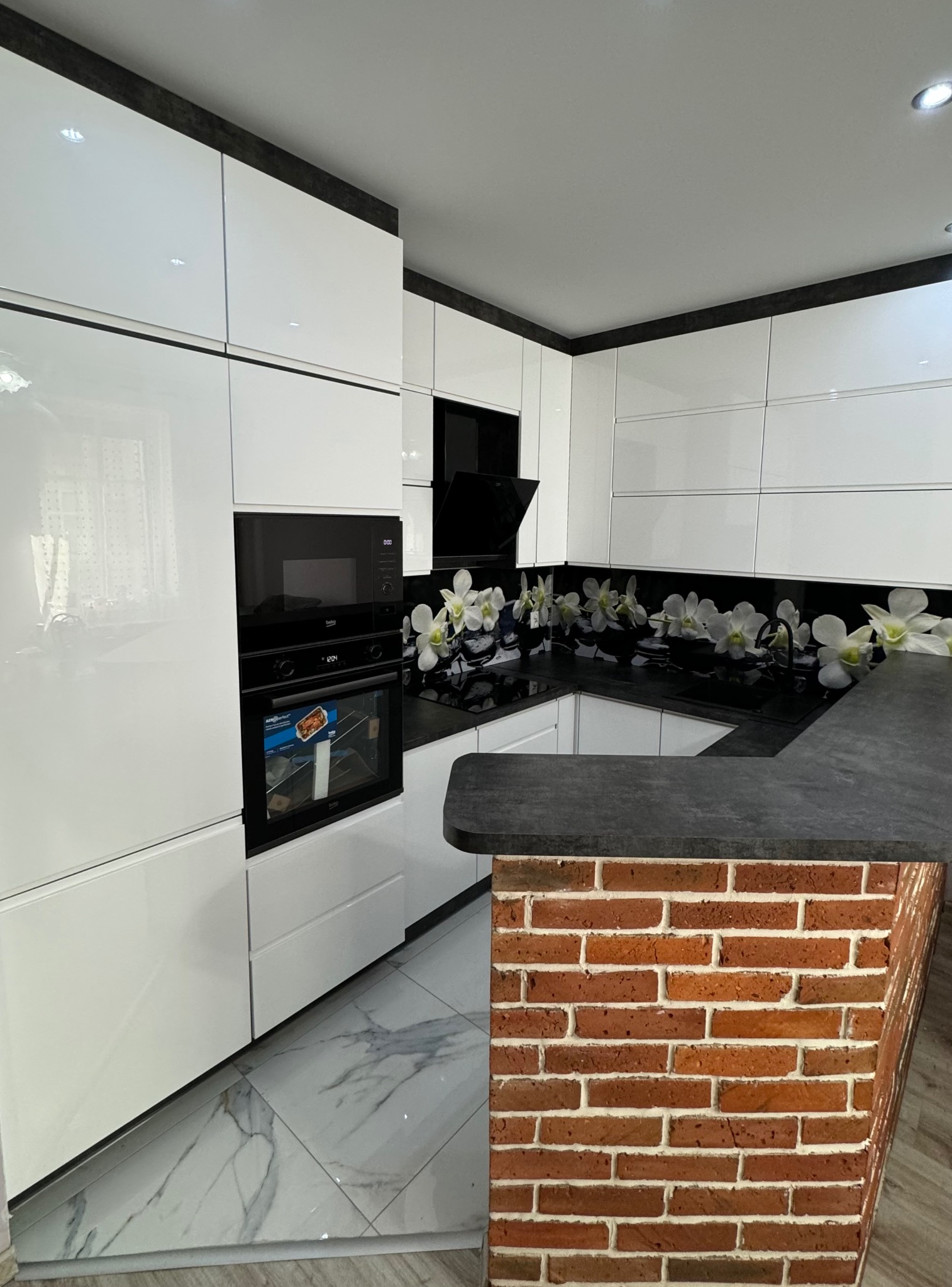 Nowoczesna kuchnia z białymi lakierowanymi frontami, blatem w kolorze ciemnego betonu i dekoracyjnym panelem szklanym z motywem białych kwiatów, uzupełniona o ceglaną zabudowę wyspy.