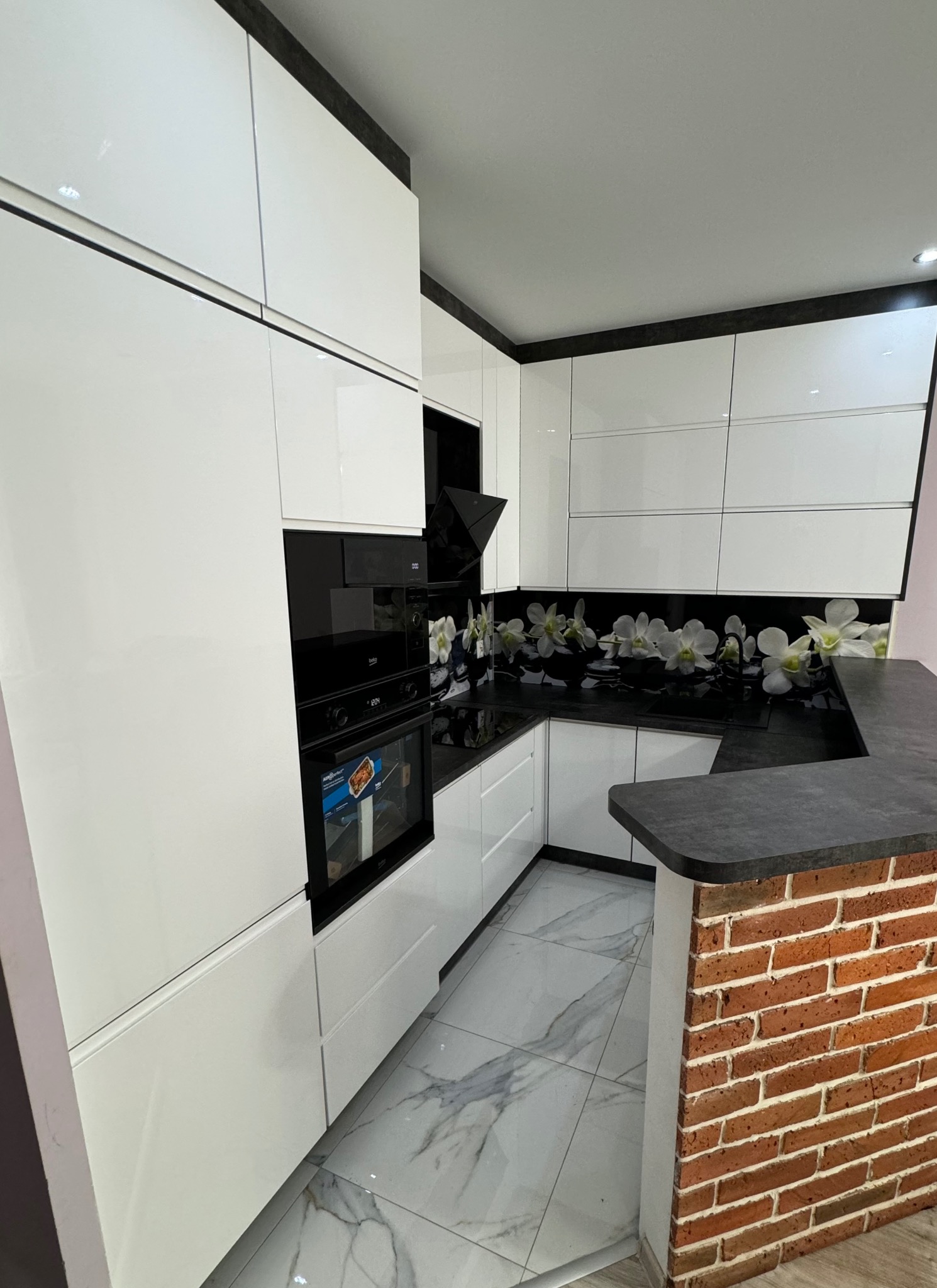 Nowoczesna kuchnia z białymi, lakierowanymi frontami, blatem w kolorze ciemnego betonu i dekoracyjnym panelem ściennym z motywem białych orchidei, uzupełniona o ceglany element barku.