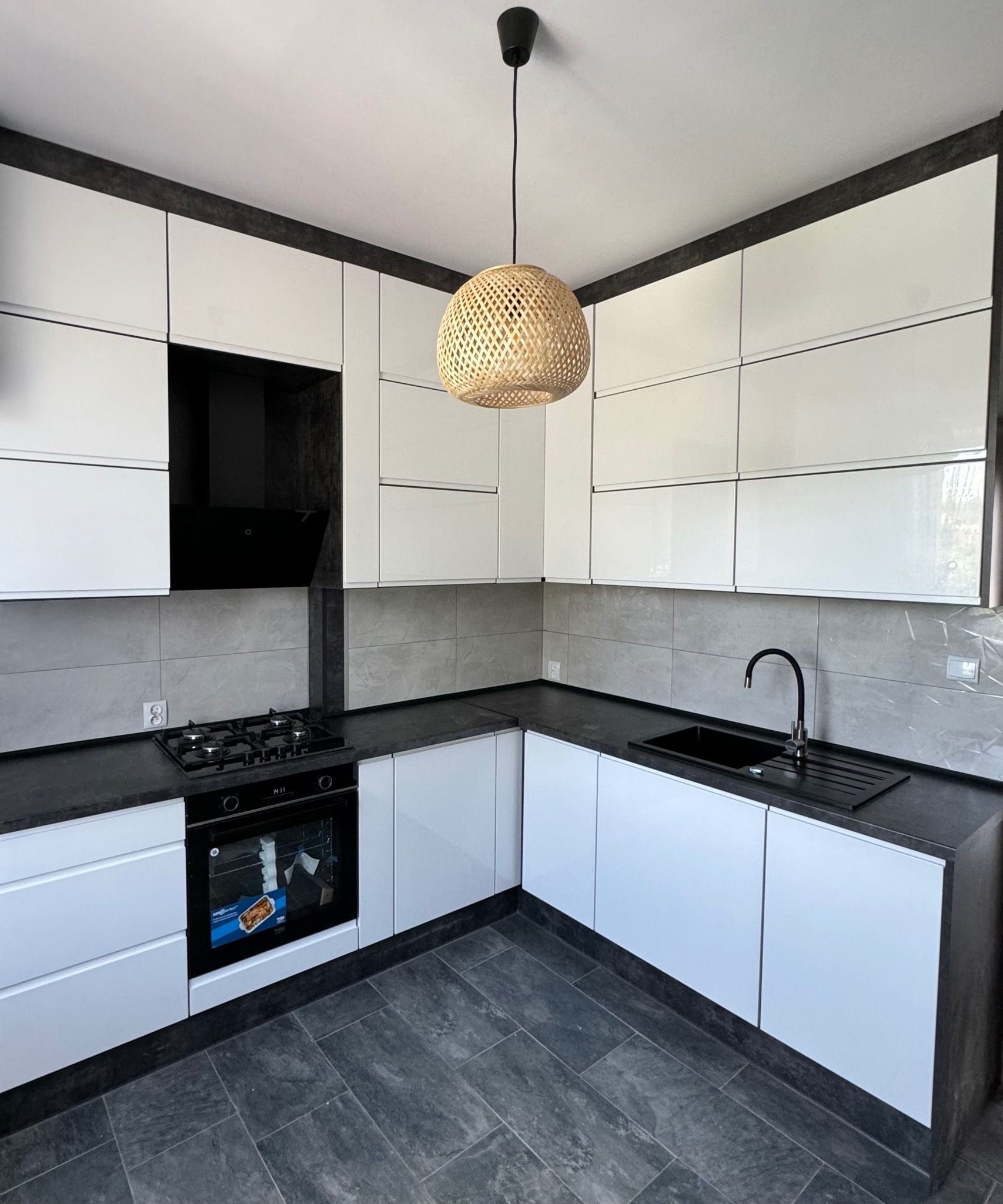 Nowoczesna kuchnia w odcieniach bieli i grafitu z lakierowanymi frontami, grafitowym blatem i płytkami imitującymi beton, oświetlona lampą z plecionki.
