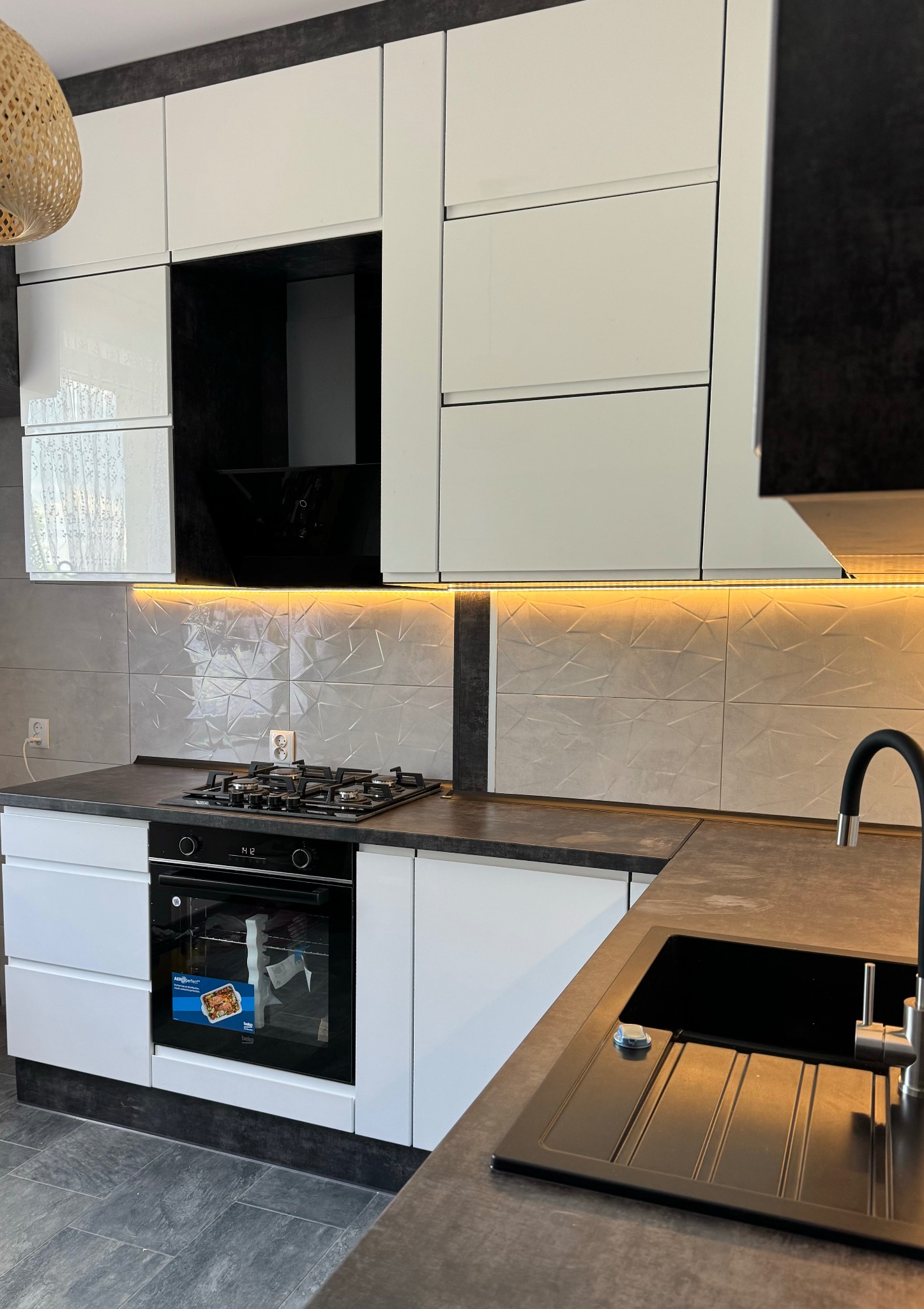 Nowoczesna kuchnia z białymi, lakierowanymi frontami szafek, blatem w kolorze ciemnego betonu, podświetleniem LED pod szafkami wiszącymi i czarną armaturą.