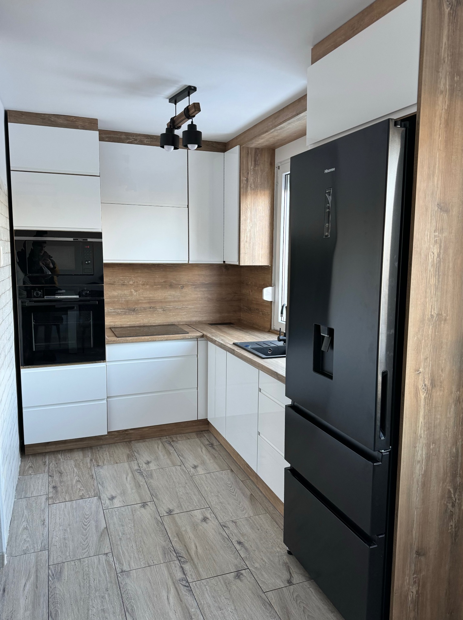 Nowoczesna kuchnia z białymi frontami, blatem i panelem ściennym w kolorze drewna retro, czarnym sprzętem AGD i drewnianą lampą sufitową.