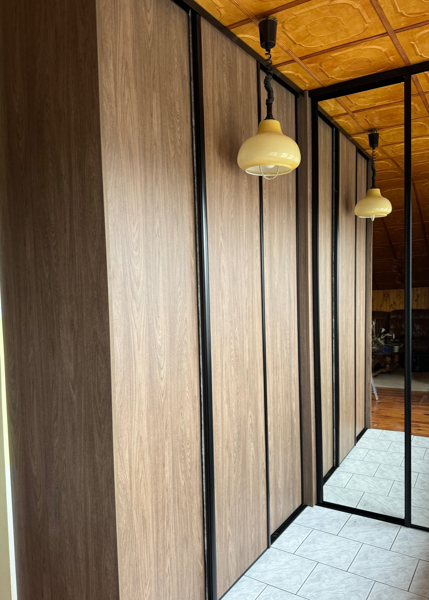 Szafa wnękowa z przesuwnymi drzwiami w odcieniach dębu sztokholm, z czarnymi profilami, widoczne odbicie wnętrza pokoju w lustrzanych drzwiach i żółta lampa wisząca.