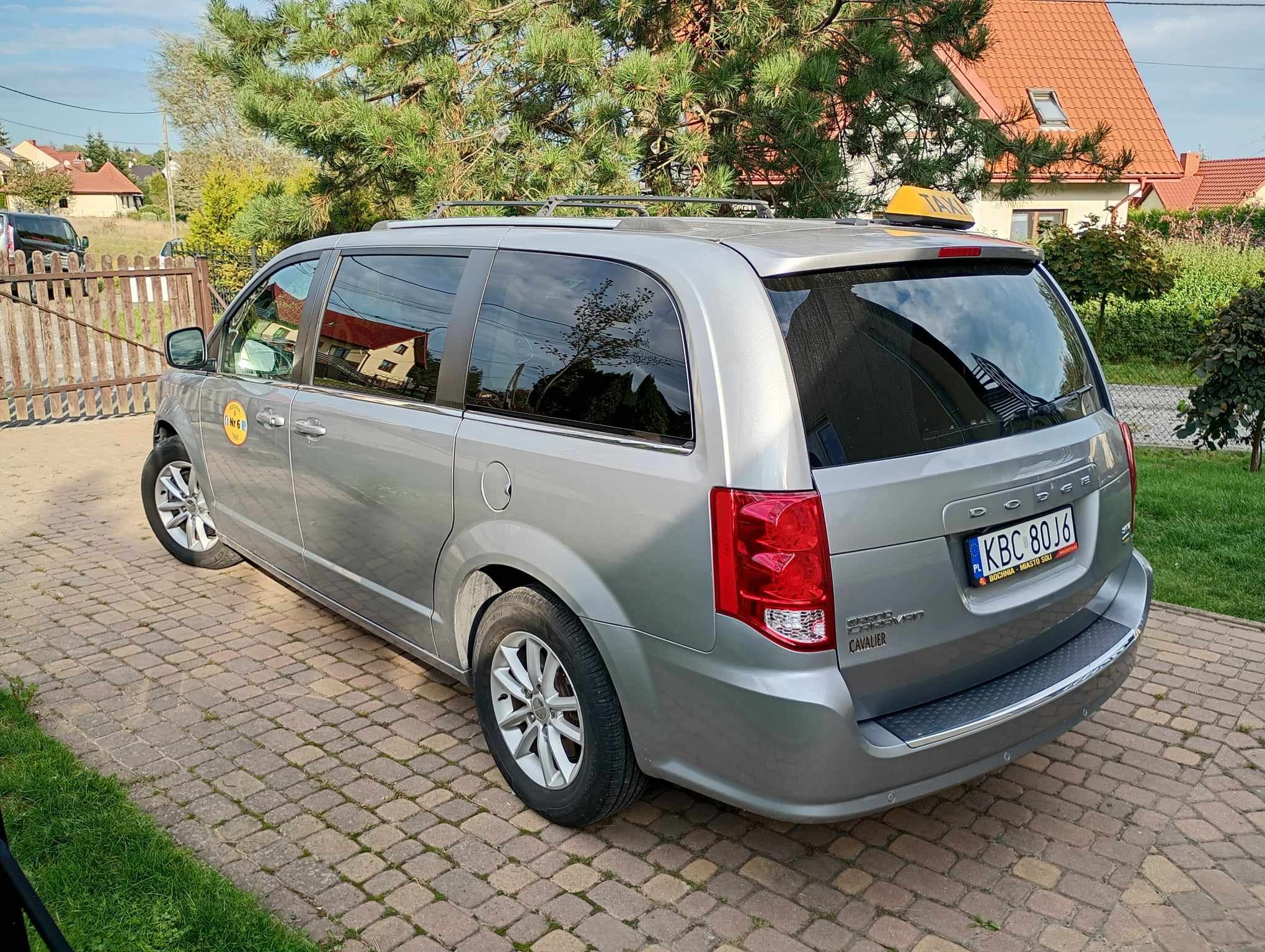 Szary van Dodge Grand Caravan z żółtym oznaczeniem Taxi na dachu, zaparkowany na brukowanej nawierzchni przed domem z czerwoną dachówką, widok z tyłu.