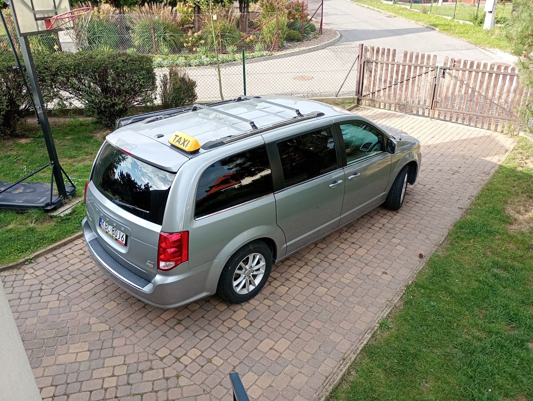 Srebrny van z żółtym znakiem Taxi na dachu, zaparkowany na brukowanym podjeździe. Widok z góry, z boku. W tle ogrodzenie i zieleń.