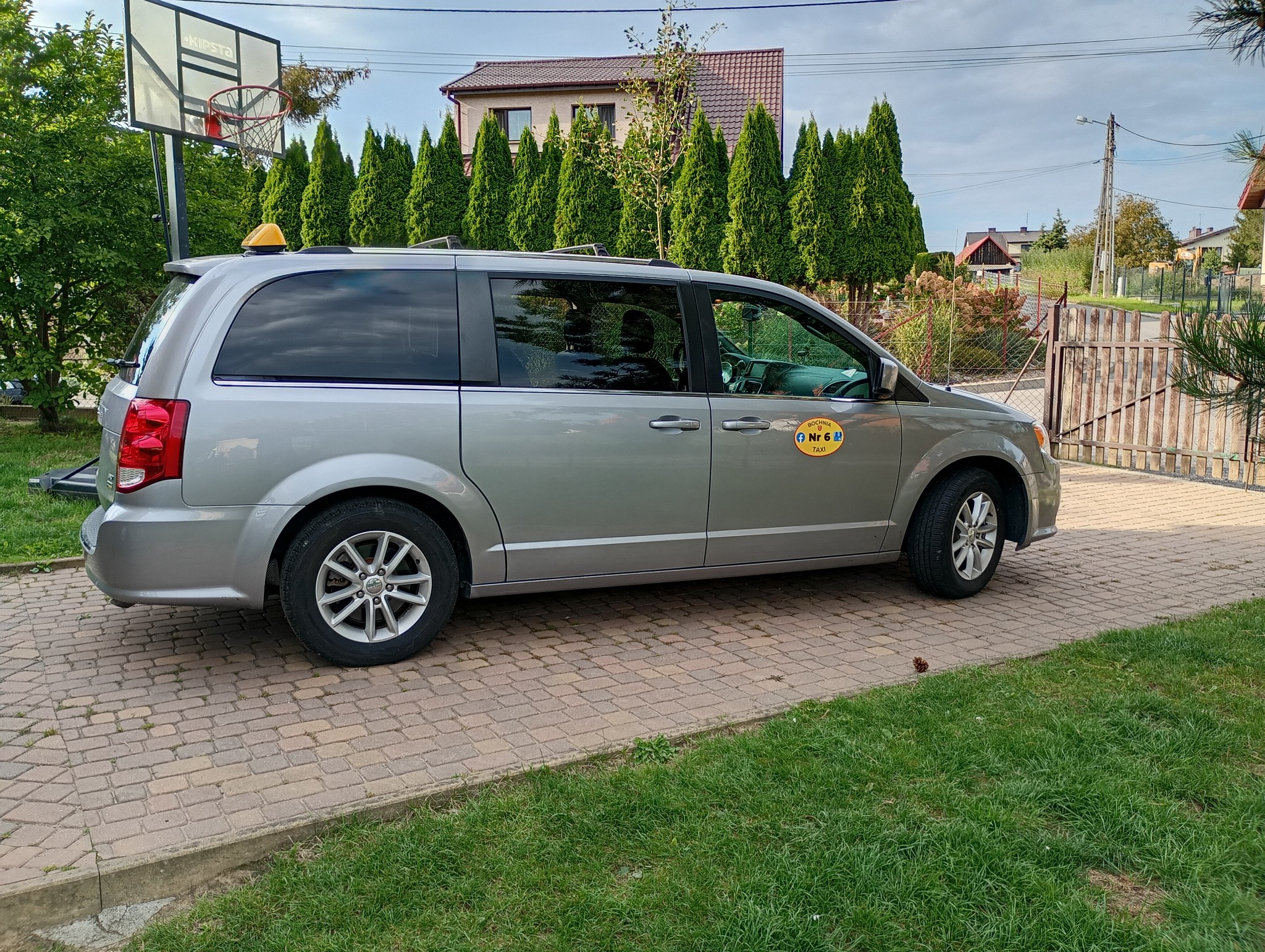 Srebrny van z żółtym oznaczeniem taxi na tle zieleni i domu. Na dachu charakterystyczny, żółty znak taxi. Widok z boku, na podjeździe z kostki brukowej.