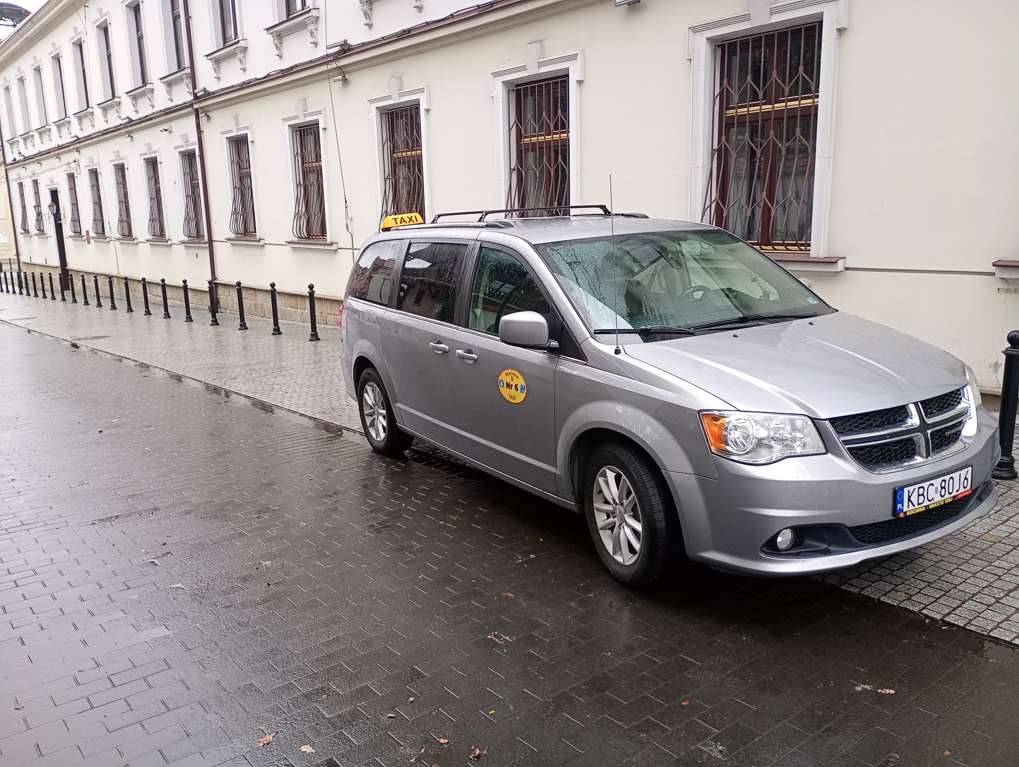Srebrny minivan Dodge z oznaczeniami taksówki Bochnia nr 6, zaparkowany na mokrym chodniku przed budynkiem z oknami z kratami. Tablica rejestracyjna KBC 80J6.