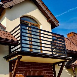 Mat&Art usługi spawalnicze Mateusz Zowisło - Czarna, pozioma barierka balkonowa z metalu, zamontowana na balkonie z ceglaną podmurówką, widok na fragment elewacji domu z brązową dachówką w słoneczny dzień.