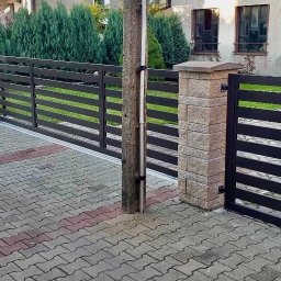Mat&Art usługi spawalnicze Mateusz Zowisło - Nowoczesne ogrodzenie metalowe z poziomymi przęsłami i słupkami z kamienia elewacyjnego, brama przesuwna i furtka, widok z zewnątrz na posesję z kostką brukową.