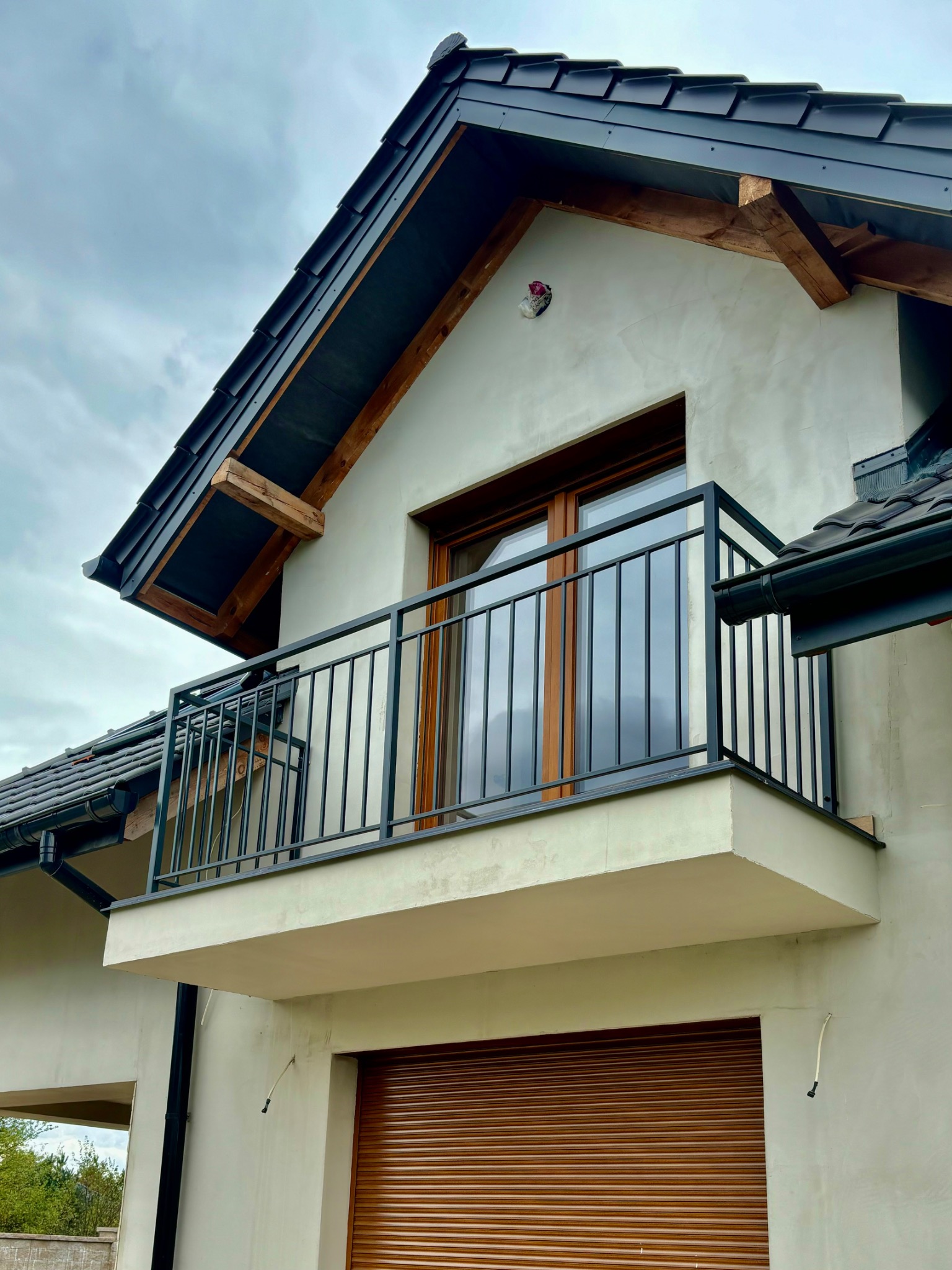 Nowoczesny balkon z czarną, metalową balustradą na tle jasnej elewacji domu z brązowymi roletami.