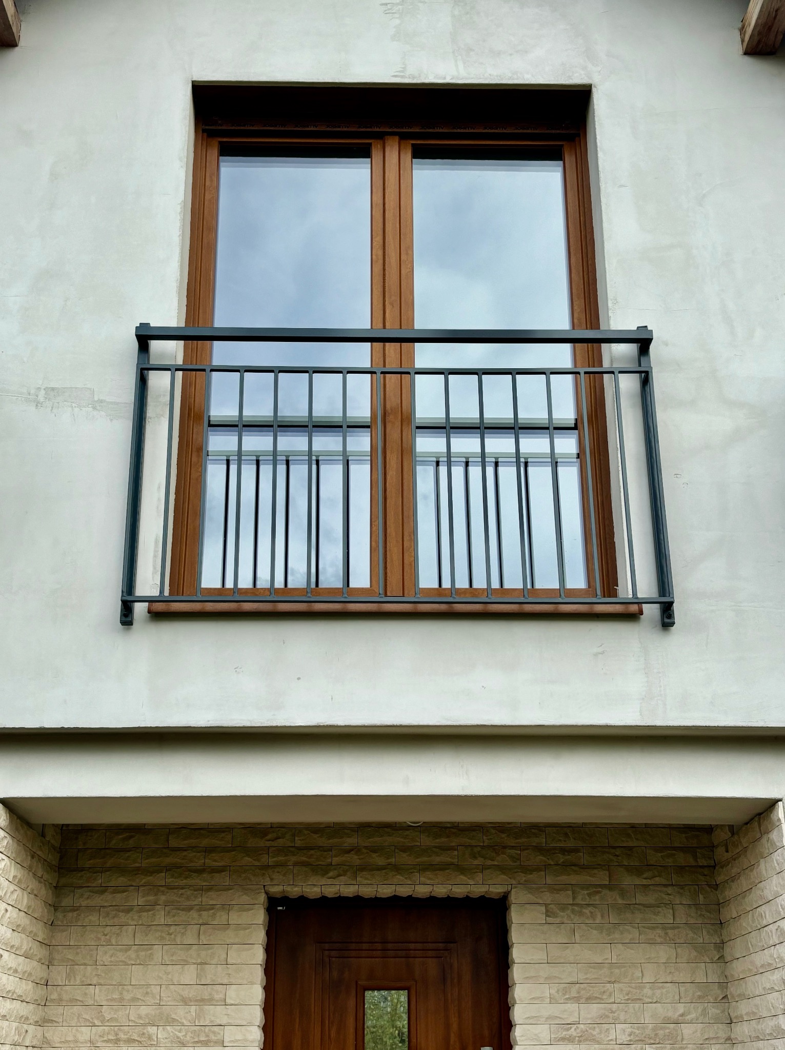 Balkon z metalową balustradą na tle szarej elewacji i brązowych ram okiennych, widok z dołu na dwuskrzydłowe okno.