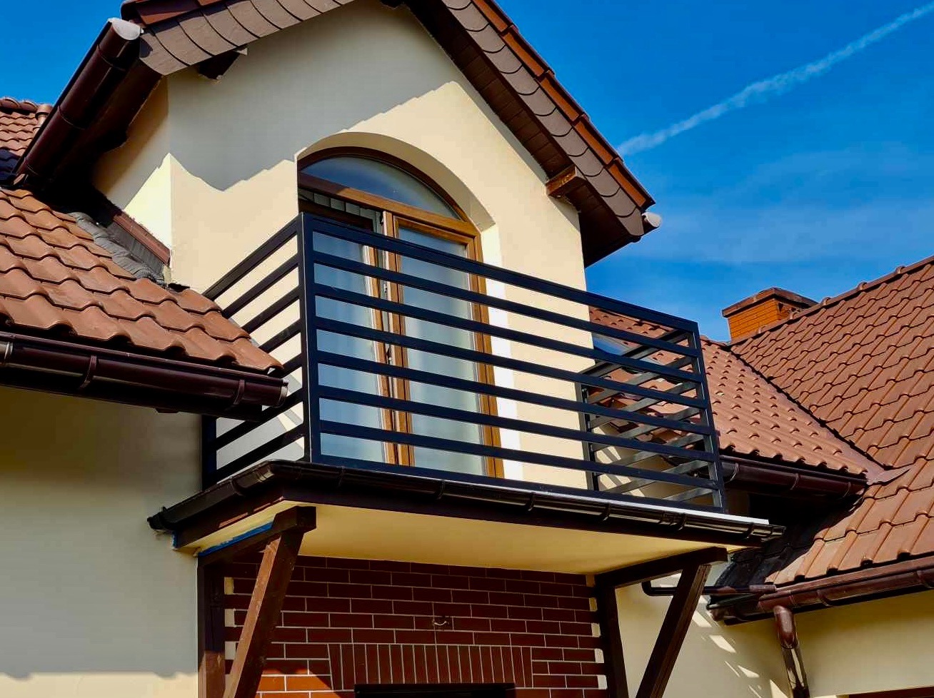 Czarna, pozioma barierka balkonowa z metalu, zamontowana na balkonie z ceglaną podmurówką, widok na fragment elewacji domu z brązową dachówką w słoneczny dzień.