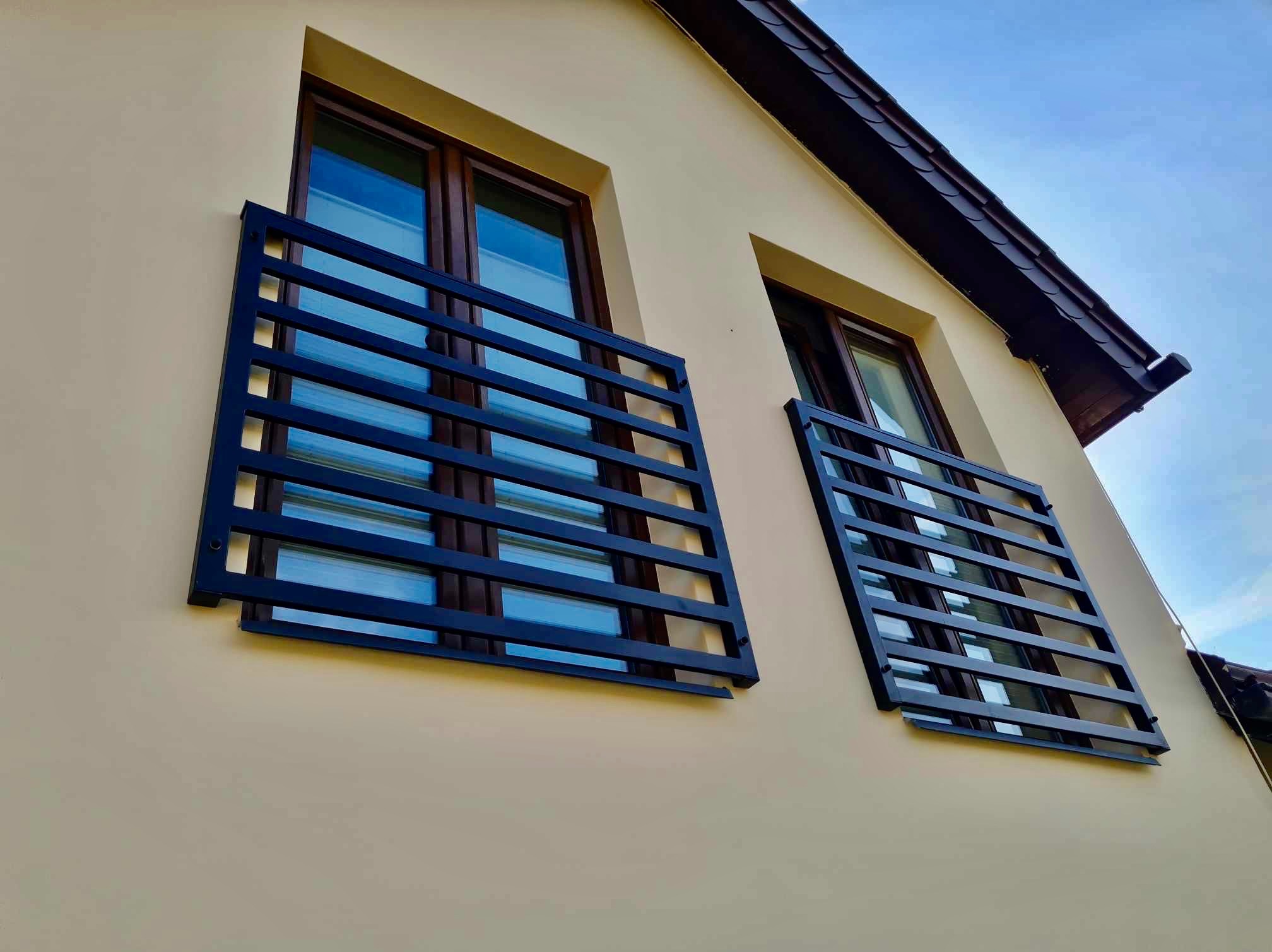 Dwa balkony z nowoczesnymi, poziomymi, metalowymi zabezpieczeniami w kolorze ciemnego granatu, zamontowane na elewacji budynku o kremowym odcieniu, widoczne z dołu pod kątem.