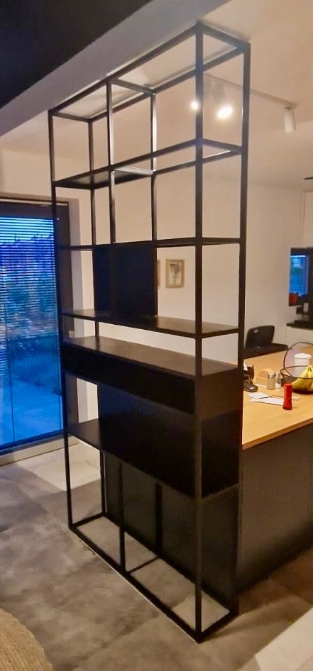 Czarna, metalowa konstrukcja regału z półkami, oddzielająca przestrzeń w nowoczesnym wnętrzu, widok na okno z żaluzjami i fragment blatu kuchennego.