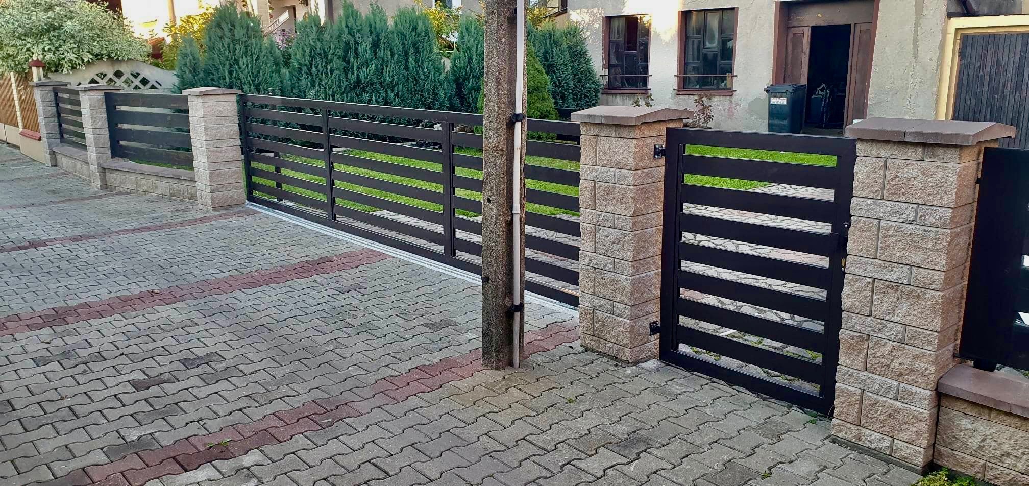 Nowoczesne ogrodzenie metalowe z poziomymi przęsłami i słupkami z kamienia elewacyjnego, brama przesuwna i furtka, widok z zewnątrz na posesję z kostką brukową.