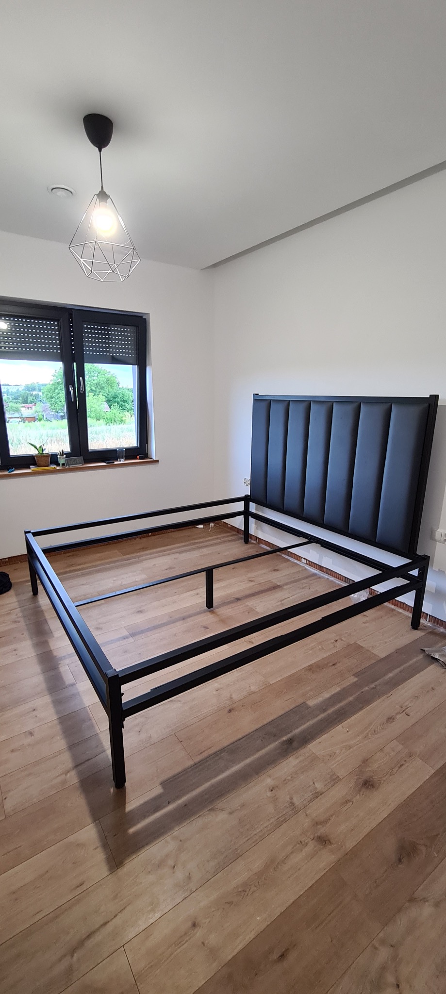 Rama metalowa łóżka z czarnym, tapicerowanym zagłówkiem w minimalistycznym wnętrzu z drewnianą podłogą i oknem z roletami.