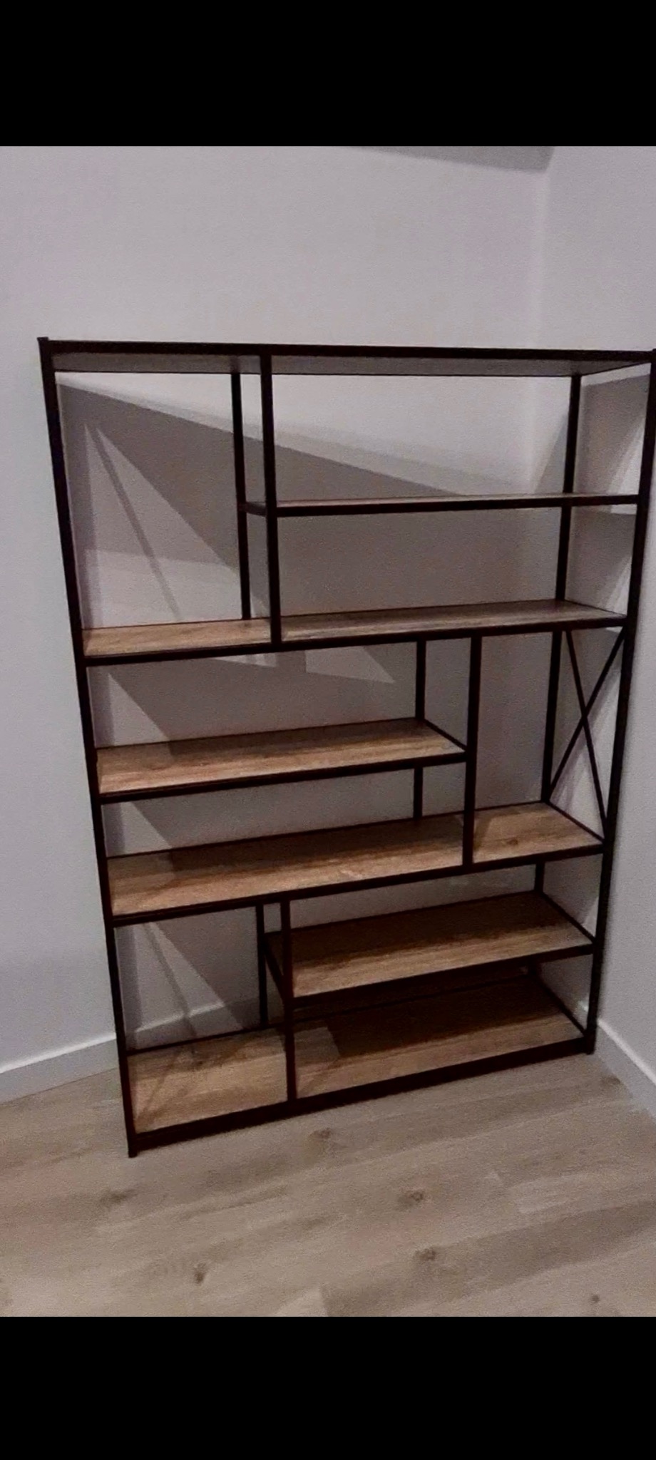 Regał metalowy z drewnianymi półkami, nowoczesny design, asymetryczny układ, ciemna rama, jasne drewno, ustawiony przy białej ścianie na podłodze imitującej drewno.