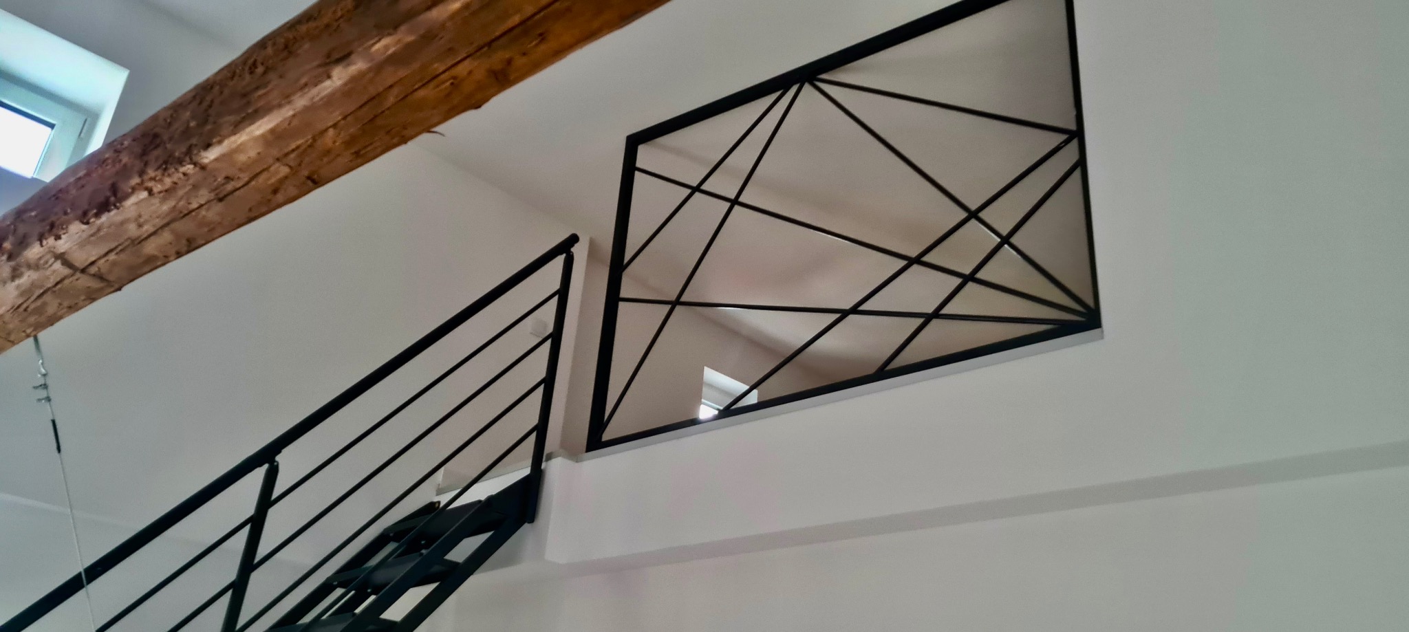 Schody z czarną, metalową konstrukcją i geometryczną balustradą wewnętrzną z widoczną drewnianą belką stropową