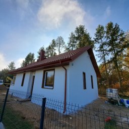 Ciesielstw Dekarstwo Artur Kolasiński - Nowo wybudowany, otynkowany dom jednorodzinny z czerwoną dachówką, rynnami i brązowymi oknami, ogrodzony siatką, z widocznym na posesji sprzętem budowlanym i drzewami w tle.