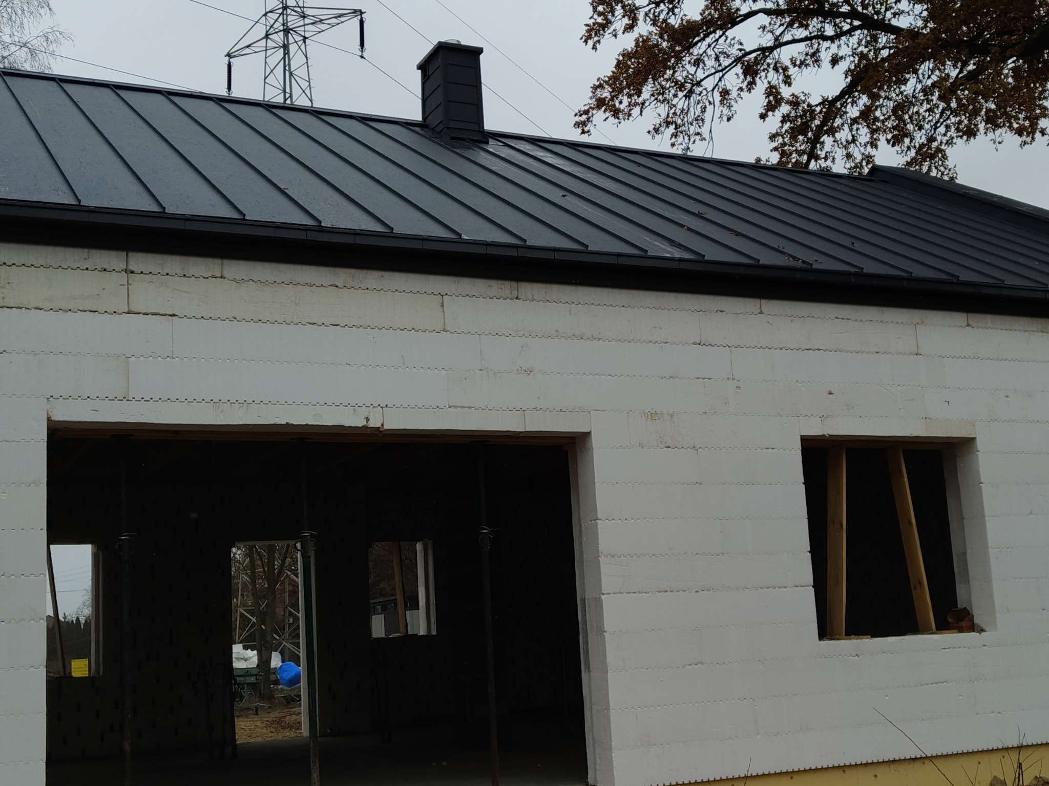 Nowy, czarny dach z blachy na budynku w trakcie budowy, widoczne okna w surowym stanie i fragment konstrukcji dachu z kominem.