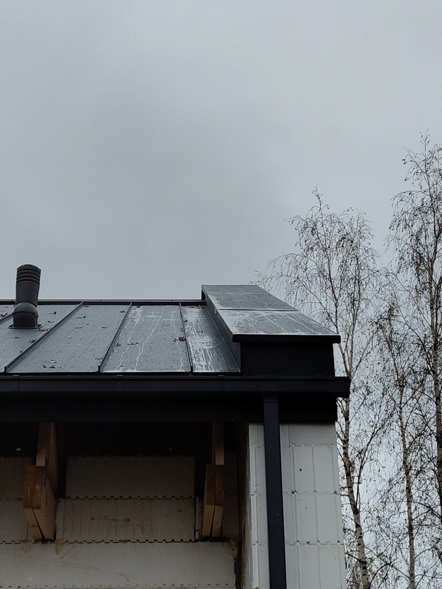 Metalowa konstrukcja dachu z kominem i rynną, widoczna na tle surowej ściany budynku z bloczków i drzew bez liści.