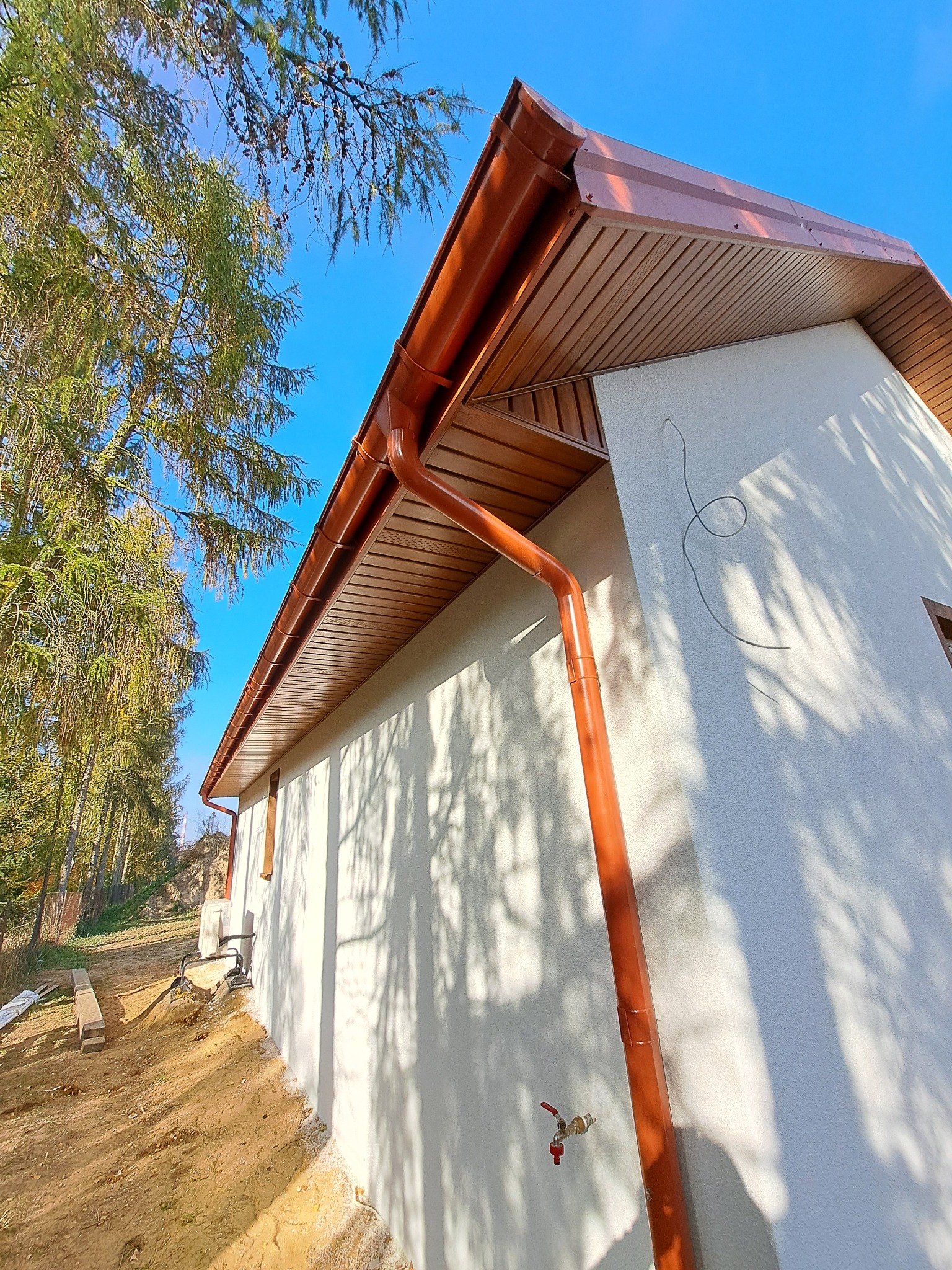 Nowoczesny dom jednorodzinny z brązowym dachem i rynnami, widok z dołu na jasnym tle nieba i drzew