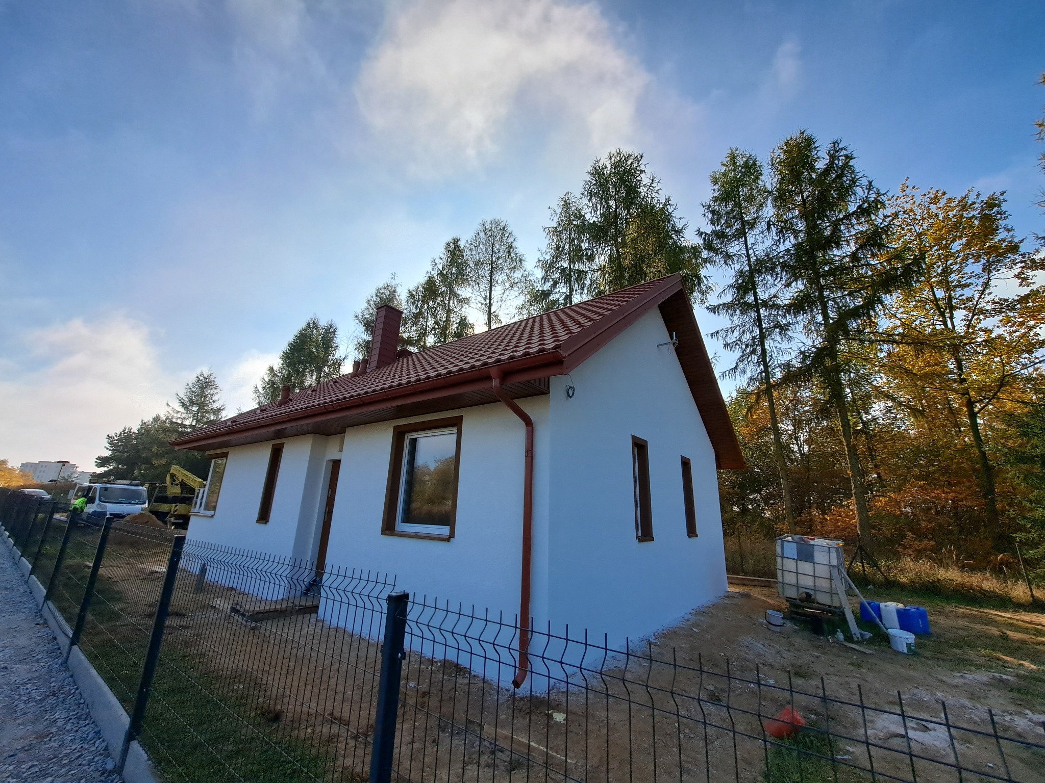 Nowo wybudowany, otynkowany dom jednorodzinny z czerwoną dachówką, rynnami i brązowymi oknami, ogrodzony siatką, z widocznym na posesji sprzętem budowlanym i drzewami w tle.