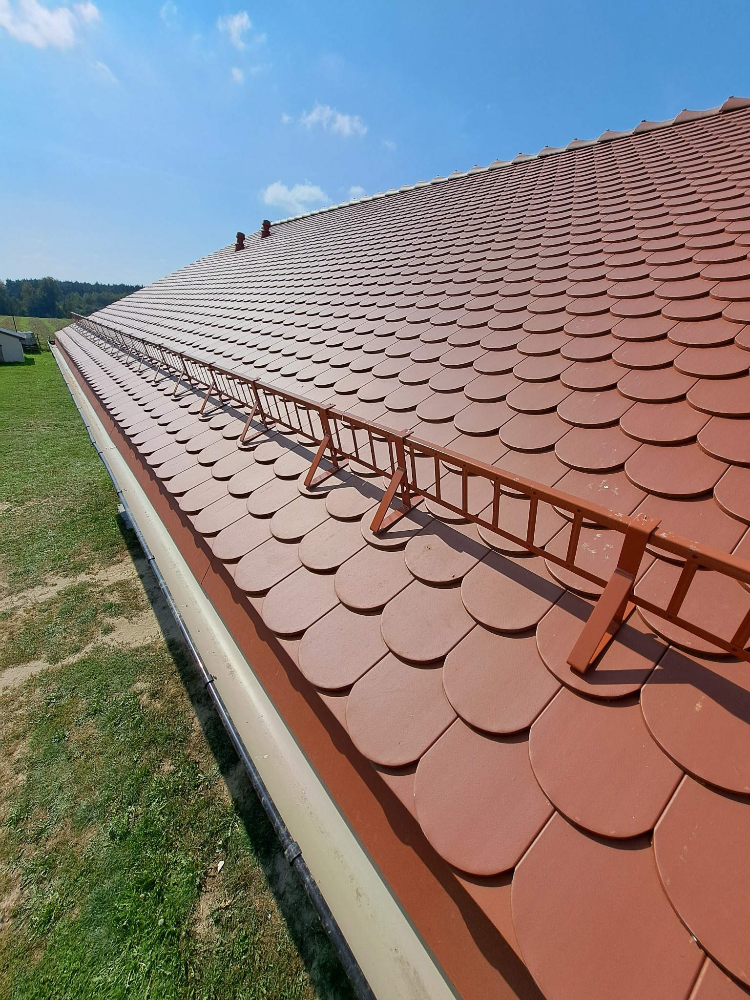 Dach pokryty czerwoną dachówką ceramiczną, zabezpieczony metalowymi barierkami śniegowymi, widok z dołu na tle błękitnego nieba.