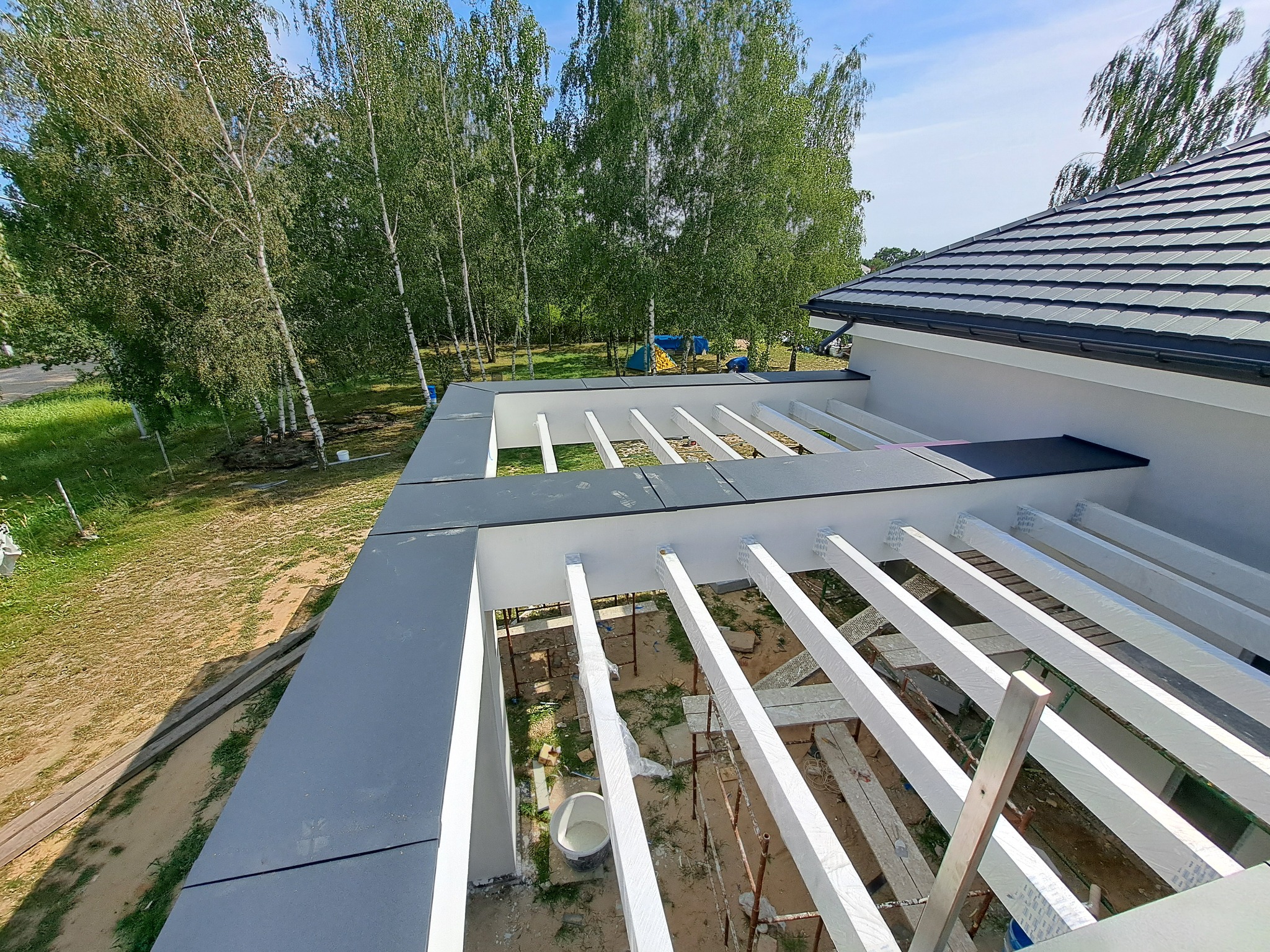 Montaż drewnianej konstrukcji pod zadaszenie na płaskim dachu, białe belki, widoczne rusztowania i wiadro z farbą, w tle brzozowy las.