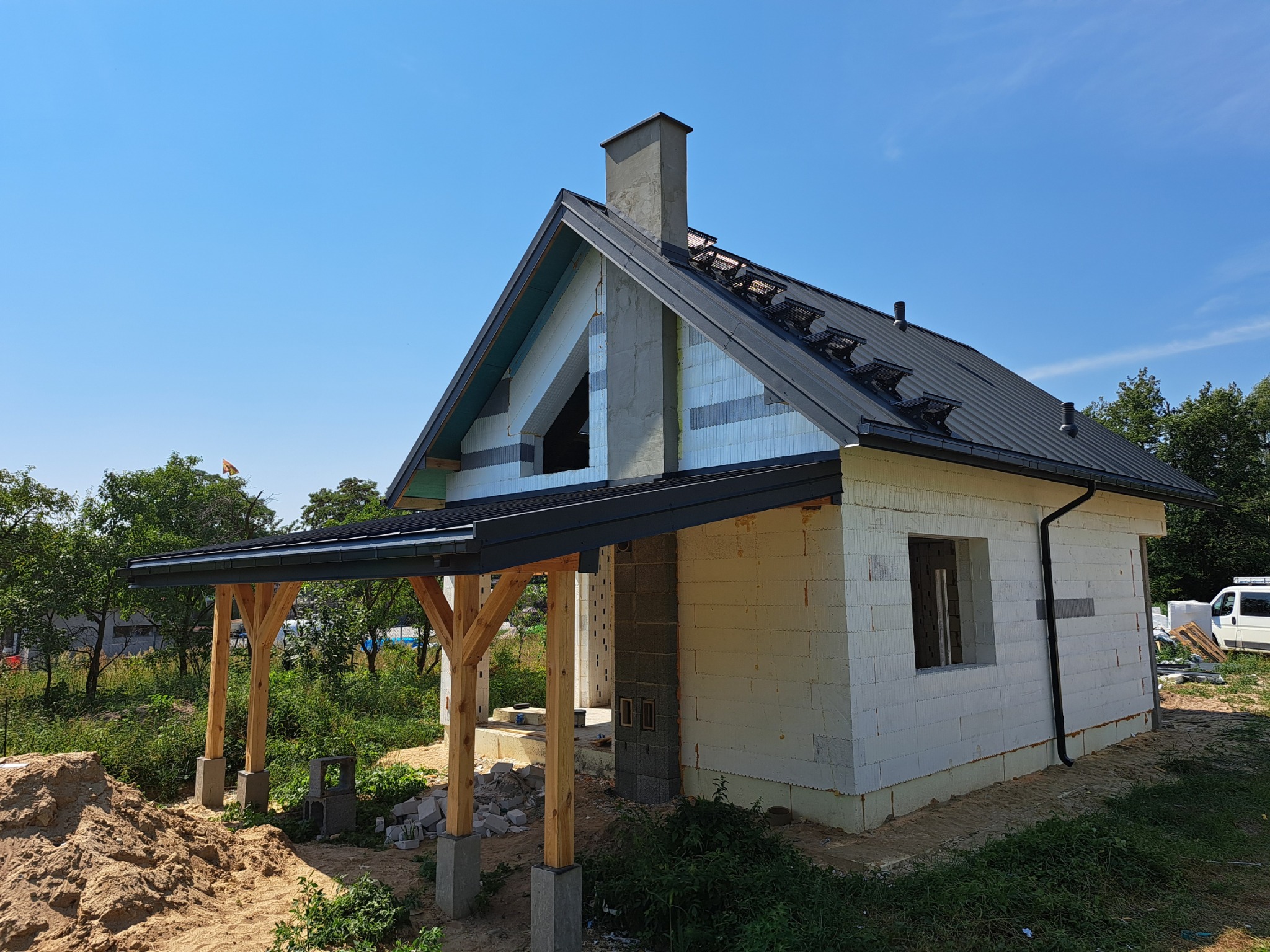 Surowy stan domu jednorodzinnego z czarnym dachem blaszanym, widoczne elementy więźby i komin, zadaszenie tarasu na drewnianych słupach, ściany z białego betonu komórkowego.