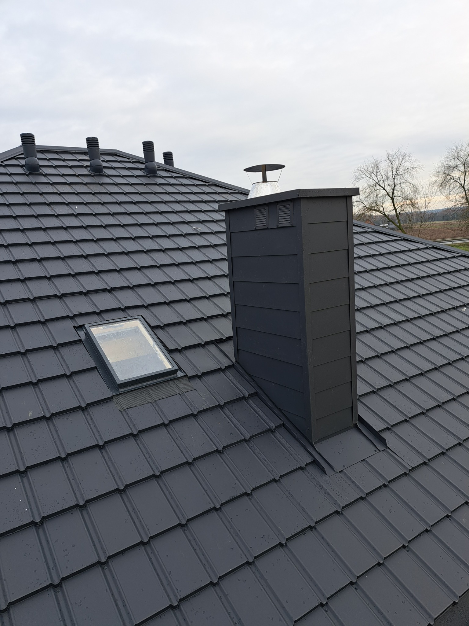 Grafitowy dach z blachodachówki z oknem dachowym i kominem wentylacyjnym, widoczne krople deszczu.