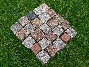 Ułożony z kostek wzór rombu na tle trawy. Kostki w odcieniach szarości, brązu i beżu, o nieregularnej fakturze. Widoczne drobne nierówności i szczeliny między kostkami.