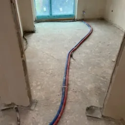 Wnętrze w trakcie remontu z ułożonymi na podłodze przewodami instalacji wodnej w kolorze niebieskim i czerwonym, widoczne surowe ściany, okno i elementy instalacji przy ścianie.