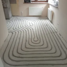 Wnętrze w trakcie montażu ogrzewania podłogowego: rury zatopione w wylewce, widoczne grzejniki na ścianach, okno i fragment wyposażenia kuchennego.