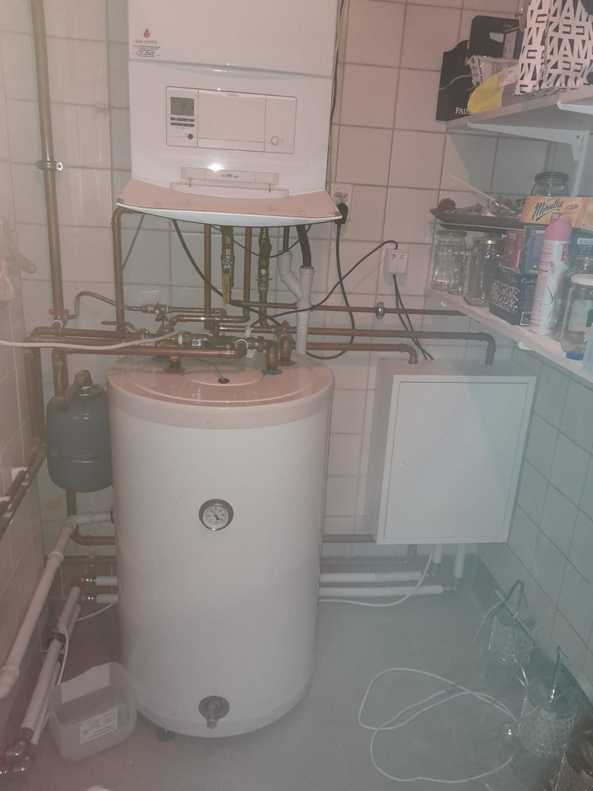 Kocioł gazowy wiszący nad zasobnikiem ciepłej wody z widocznymi rurami miedzianymi w pomieszczeniu z wykafelkowanymi ścianami i półką z artykułami spożywczymi.