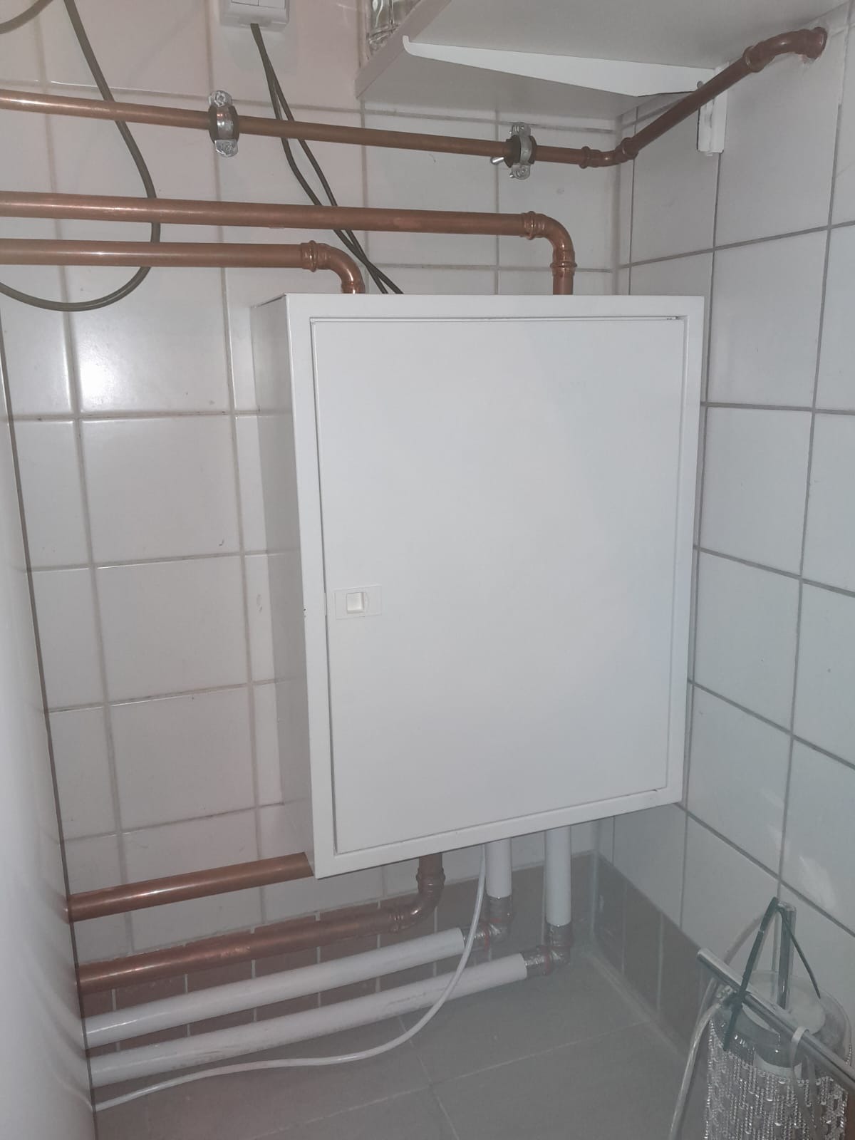 Biała szafka z instalacją hydrauliczną w tle, rury miedziane i plastikowe, białe płytki na ścianach.