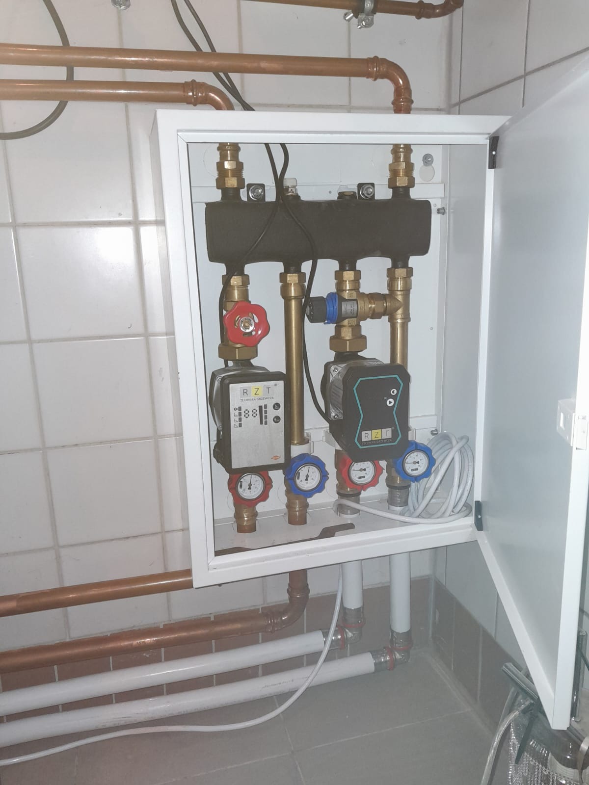 Rozdzielacz ogrzewania podłogowego z dwoma pompami obiegowymi RZT w białej szafce, widoczne miedziane rury instalacji grzewczej i manometry z niebieskimi i czerwonymi wskazówkami.