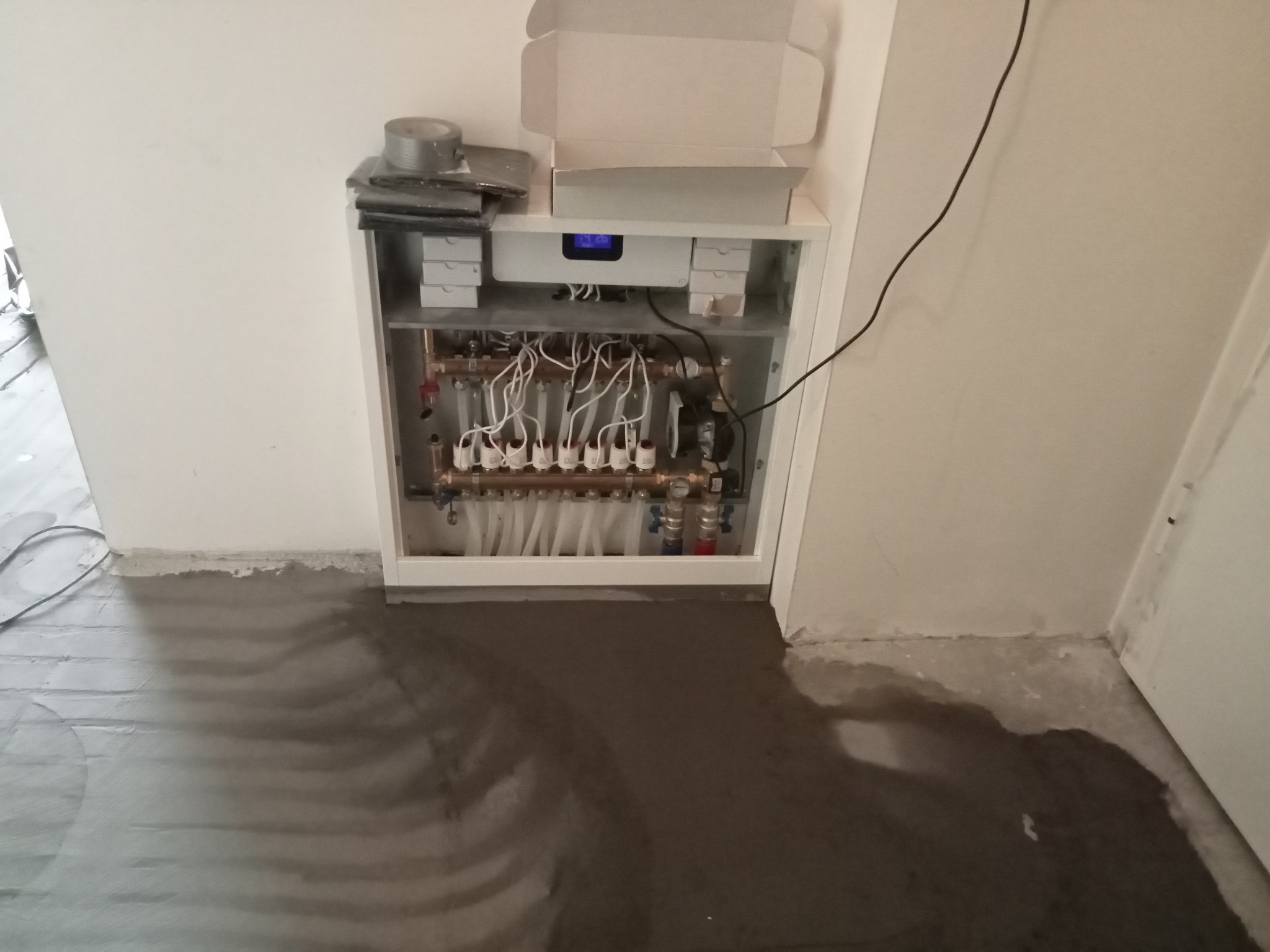 Rozdzielacz ogrzewania podłogowego w szafce, widoczne rury, zawory, kable, na górze szafki leżą materiały instalacyjne, na podłodze wylewka samopoziomująca.