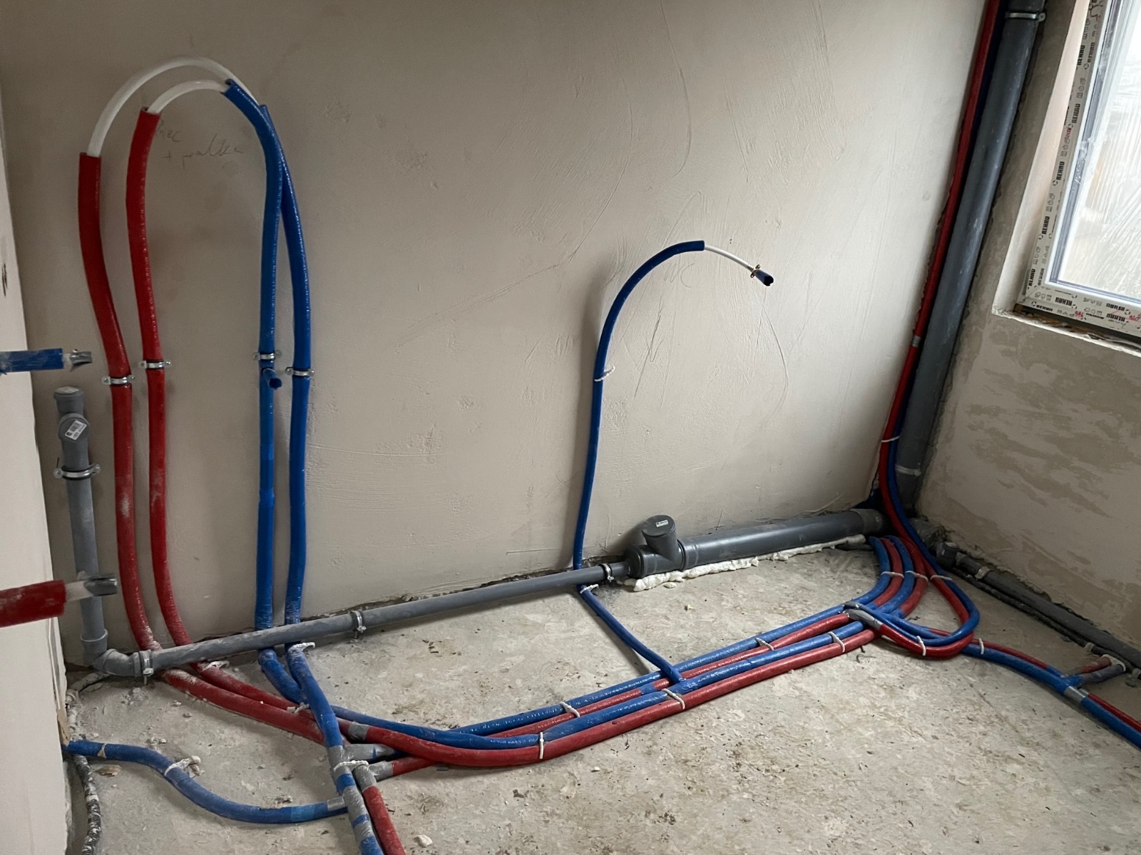 Nowa instalacja wodna i grzewcza w surowym wnętrzu, z widocznymi rurami PEX w kolorach czerwonym i niebieskim, prowadzącymi wzdłuż ścian i podłogi, przed wylewką.