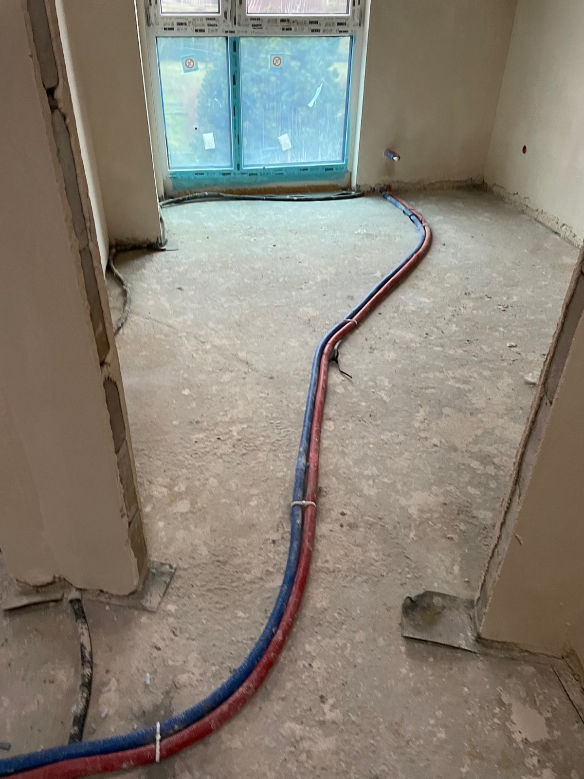 Wnętrze w trakcie remontu z ułożonymi na podłodze przewodami instalacji wodnej w kolorze niebieskim i czerwonym, widoczne surowe ściany, okno i elementy instalacji przy ścianie.