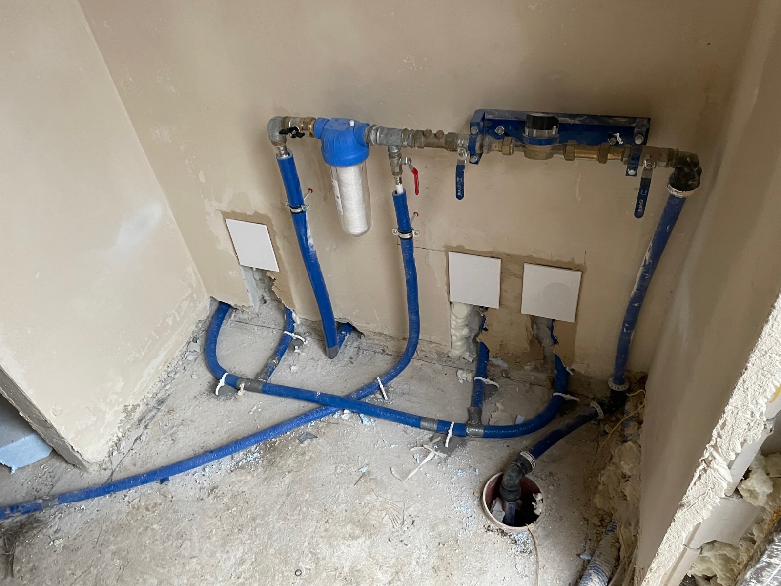 Fragment instalacji hydraulicznej w trakcie przeróbki: niebieskie rury PEX prowadzone w bruzdach w ścianie, filtr wody z przezroczystym korpusem, zawory i złączki mosiężne, ślady po kuciu w ścianie...