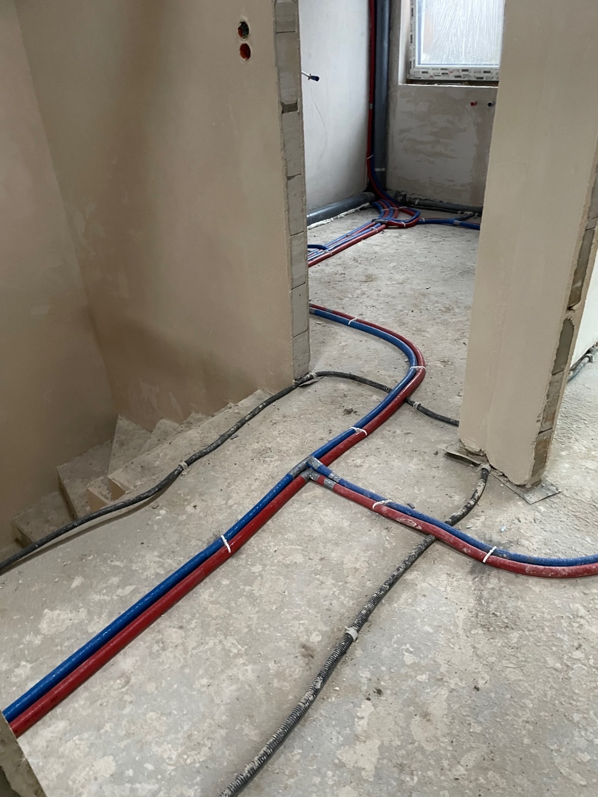 Surowe wnętrze w trakcie remontu z widocznymi, nowymi rurami instalacji wodnej w kolorach czerwonym i niebieskim, zabezpieczonymi białymi opaskami, oraz czarnym kablem elektrycznym biegnącym po...