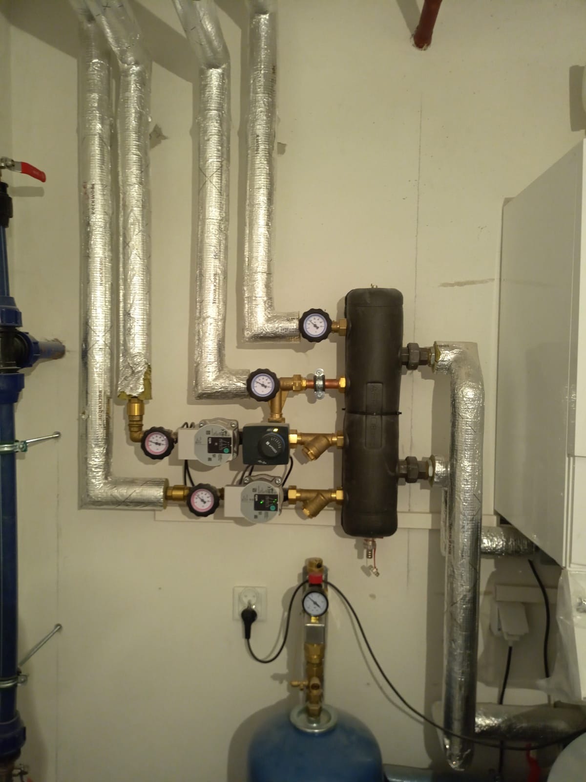 Złożona instalacja grzewcza z kotłem, zbiornikiem wyrównawczym i rurami w izolacji aluminiowej, manometry wskazują ciśnienie w systemie.