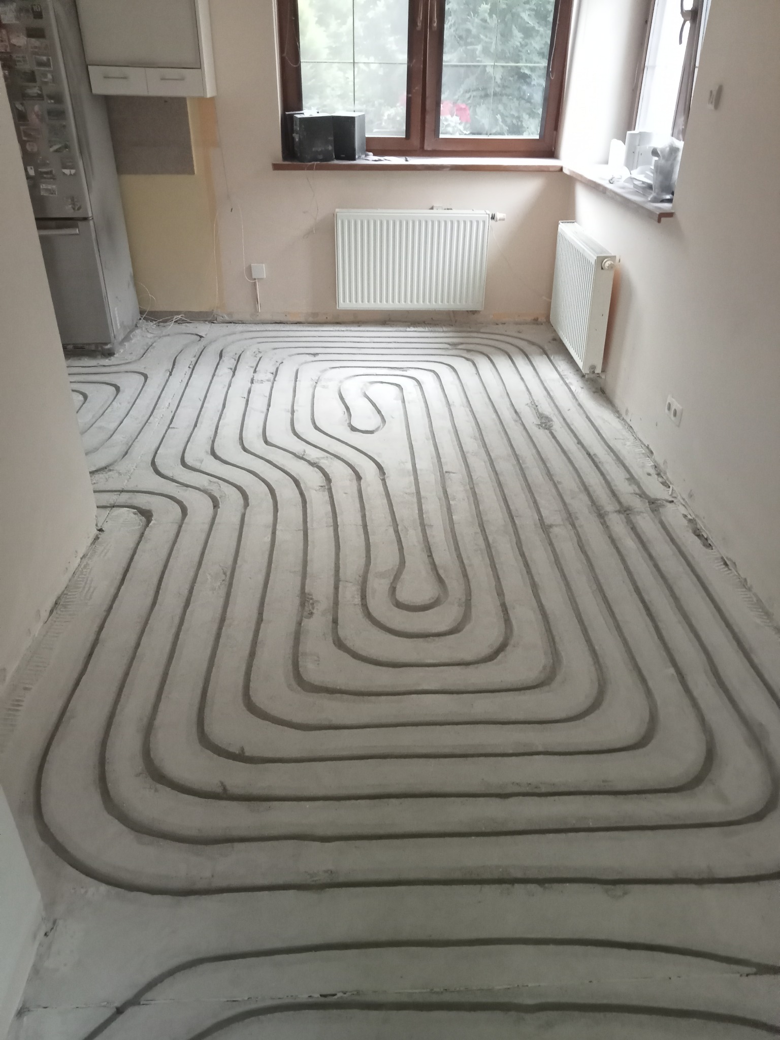 Wnętrze w trakcie montażu ogrzewania podłogowego: rury zatopione w wylewce, widoczne grzejniki na ścianach, okno i fragment wyposażenia kuchennego.
