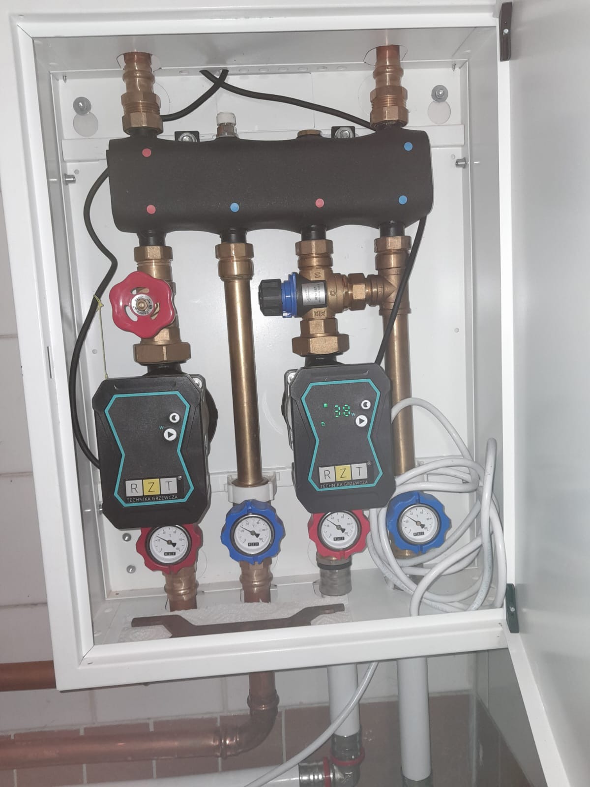 Rozdzielacz instalacji grzewczej w białej szafce, widoczne pompy obiegowe RZT, manometry z czerwonymi i niebieskimi oznaczeniami, rury miedziane i zawory.