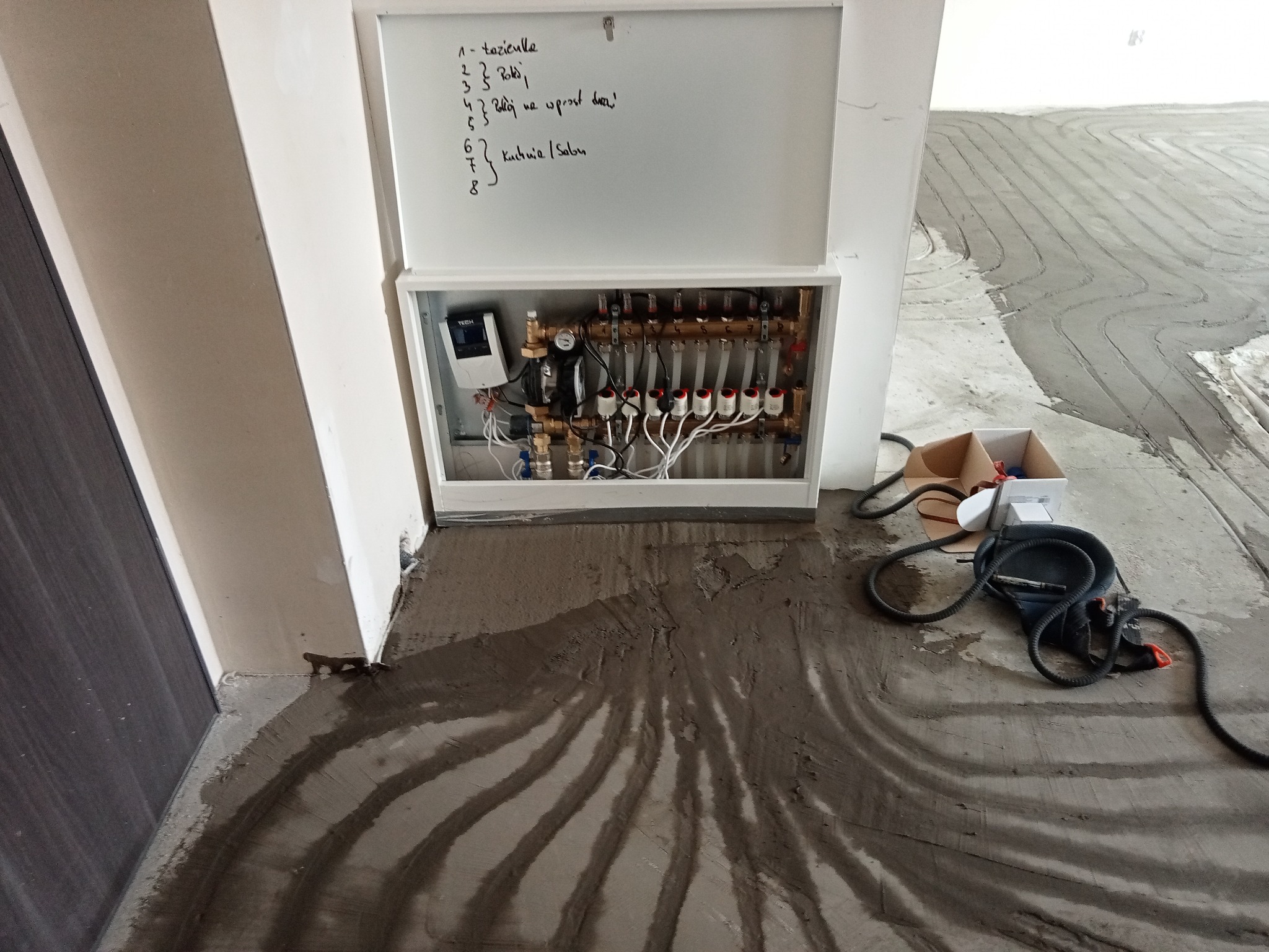 Rozdzielacz ogrzewania podłogowego w szafce z listą pomieszczeń zapisaną na drzwiczkach, widoczne rury instalacji w betonie, narzędzia obok.