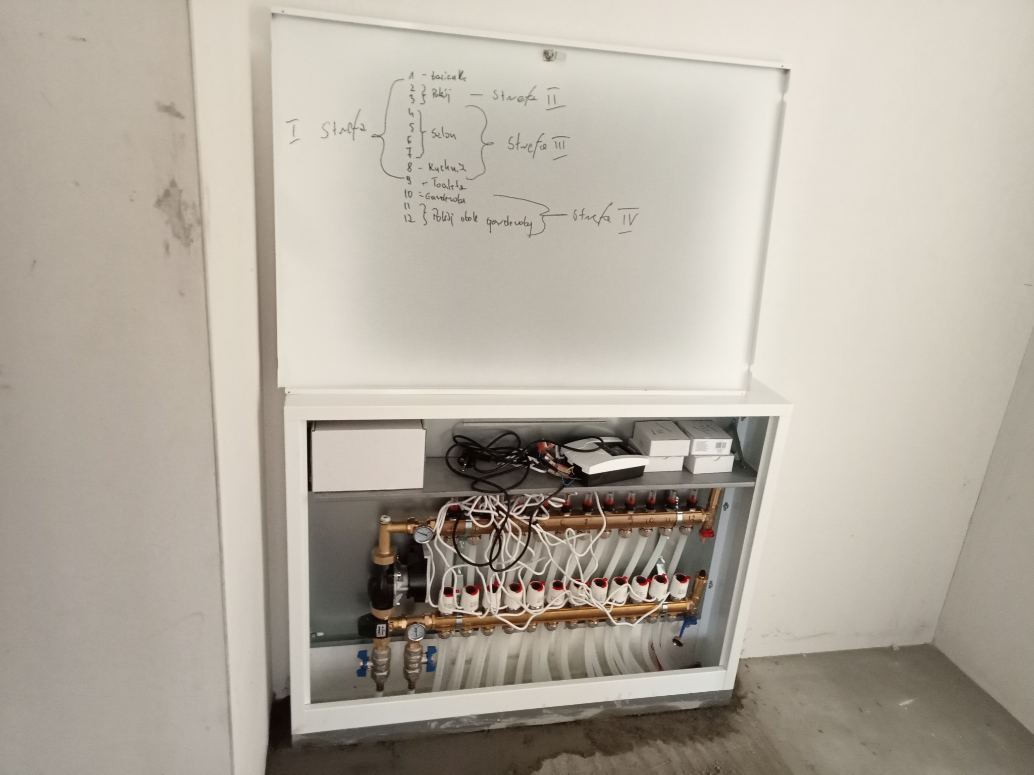 Rozdzielacz instalacji grzewczej w białej obudowie z widocznymi rurami, manometrami i zaworami, nad nim biała tablica z odręcznymi notatkami o strefach pomieszczeń.