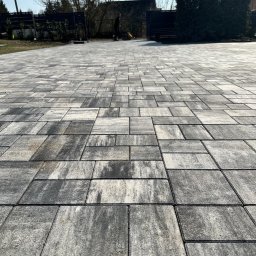 Artbruk - Ułożona szara kostka brukowa na podjeździe, widok z niskiej perspektywy, na tle domu z ciemnym ogrodzeniem i sylwetką osoby w oddali.